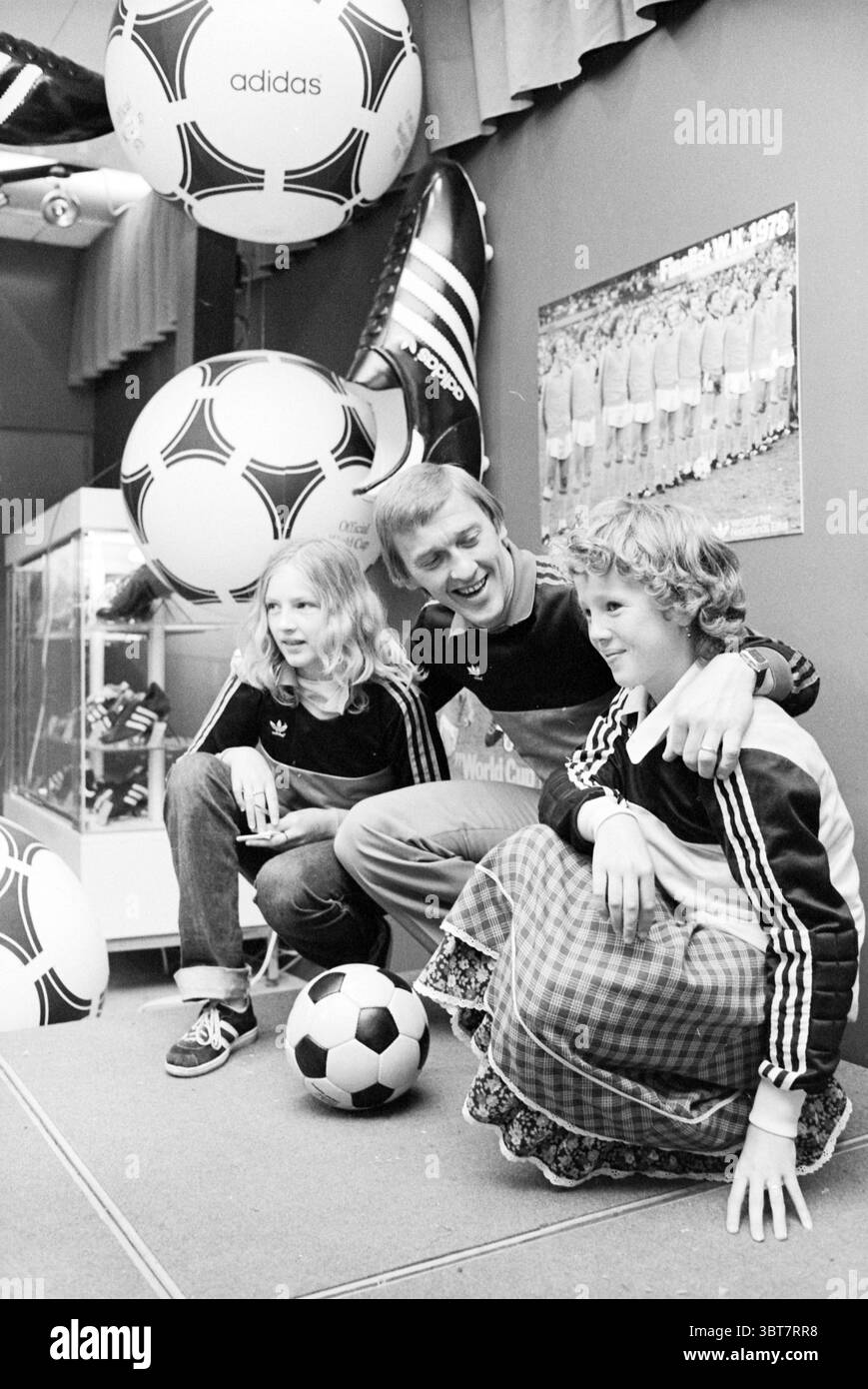 PIM Doesburg presso V&D' People Sports Shops and Kiosks, Whizgle News, Dutch Desk, Paesi Bassi, 1950 - 2000 il 12-09-1979. Questi argomenti sono mostrati nell'immagine. In un ambiente interno vivace, due bambini sono seduti accanto a un uomo adulto, tutti indossando un abbigliamento sportivo scuro abbinato a righe bianche. I bambini hanno i capelli lunghi e ondulati ed emettono un senso di eccitazione, sorridendo brillantemente mentre interagiscono con l'uomo. L'uomo, mostrando un'espressione calda, si appoggia verso di loro, migliorando l'atmosfera allegra. Intorno a loro ci sono grandi palline da calcio sospese dal soffitto, aggiungendo un playfu Foto Stock