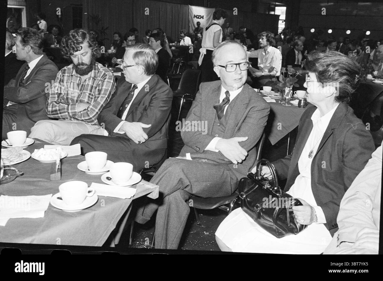 Ricevimento 100 anni di NZH Noord Zuid Hollandse Vervoersmaatschappij N.V. NZ, Whizgle News, Dutch Desk, Paesi Bassi, 1950 - 2000 il 18-05-1981. Questi sono gli elementi dell'immagine. La scena si snoda in un vivace ambiente interno, caratterizzato da grandi tavoli drappeggiati in tovaglie scure. Attorno ai tavoli vi è un gruppo eterogeneo di individui, ciascuno impegnato in conversazione o contemplazione. A sinistra, un uomo vestito in modo informale con una barba cespugliosa e una camicia a quadri si appoggia sulla sedia, trasudando un comportamento rilassato. Accanto a lui, un individuo vestito più formalmente con un vestito sembra essere profondo Foto Stock