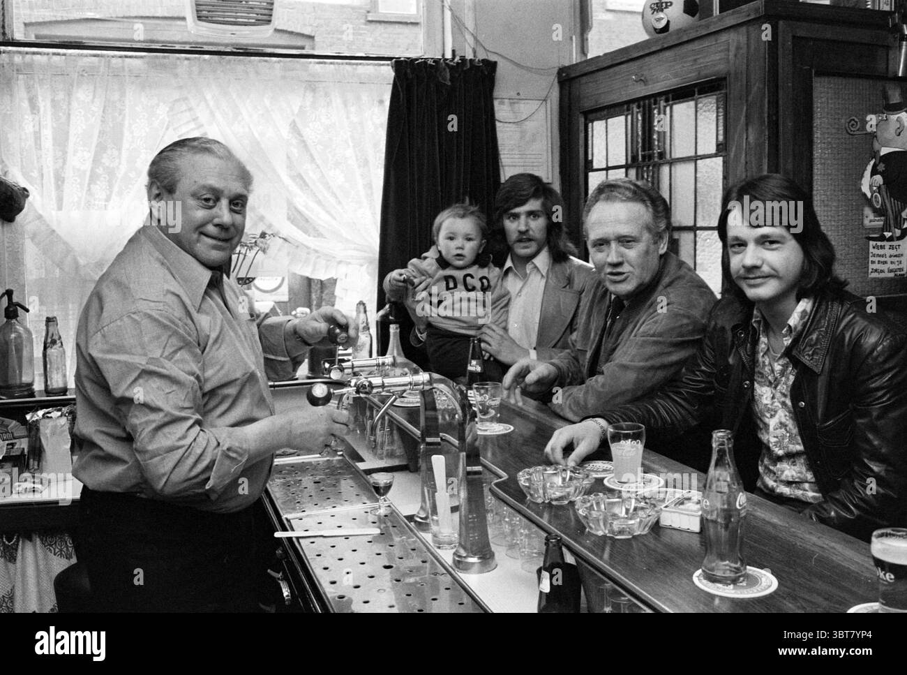 DCO in café Koornstra Café Football, Whizgle News, Dutch Desk, Paesi Bassi, 1950 - 2000 il 16-04-1976. Questi argomenti sono mostrati nell'immagine. La scena si sviluppa in un accogliente ambiente bar, caratterizzato da un'atmosfera calda e invitante. Un bancone bar in legno si estende in primo piano, lucidato per brillare, con una serie di bicchieri e bottiglie di bibite analcoliche. Dietro il bar, un uomo con un atteggiamento amichevole, rivestito di una camicia di colore chiaro, si erge attivamente, coinvolgendo i clienti. Ha un sorriso avvicinabile e probabilmente sta versando un drink. Accanto a lui, un bambino piccolo è annidato tra le braccia di un m Foto Stock