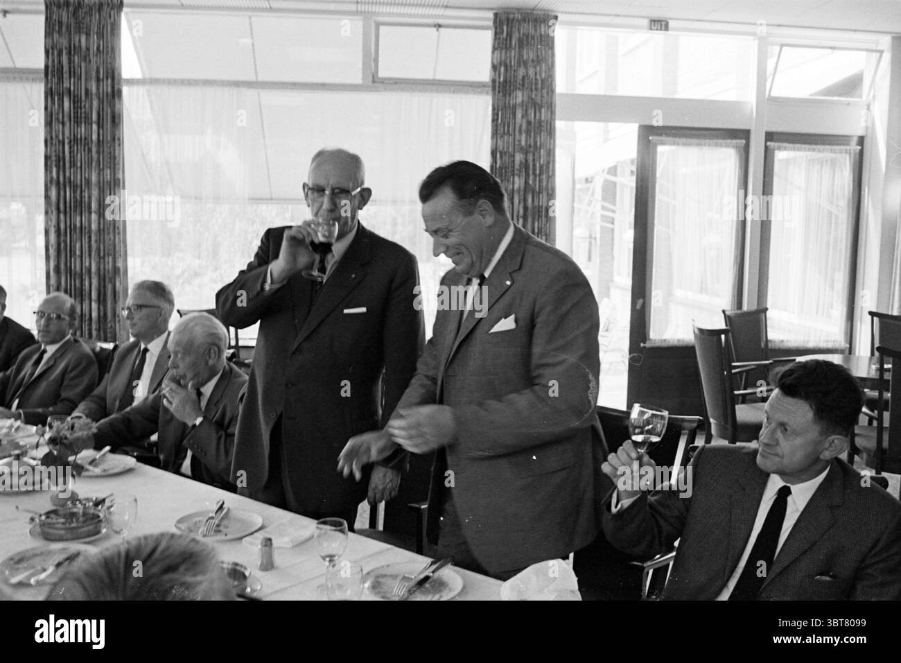 Trasferimento del nuovo autobus N.Z.H. a N.S. Noord Zuid Hollandse Vervoersmaatschappij N.V. NZ, Whizgle News, Dutch Desk, Paesi Bassi, 1950 - 2000 il 25-08-1967. Questi argomenti vengono visualizzati nell'immagine. La scena cattura un incontro di uomini ben vestiti seduti a un lungo tavolo da pranzo, con tovaglie bianche e posate ordinate. Ogni individuo trasuda un senso di formalità e cameratismo, con alcuni bicchieri da vino in alto in un brindisi. Gli uomini variano in età, indossando abiti che riflettono gli stili di un'epoca passata, con tonalità più scure che dominano la palette. La camera dispone di grandi finestre che consentono di Foto Stock