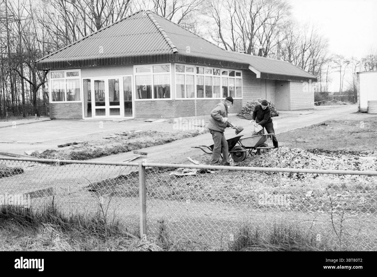 Clubhouse Cycling Association [De] Kampioen Spaarnwoude Cycling, Whizgle News, Dutch Desk, Paesi Bassi, 1950 - 2000 il 01-03-1989. L'immagine include questi argomenti. La scena raffigura un cantiere adiacente a un edificio moderno con un caratteristico tetto in piastrelle rosse e grandi finestre in vetro che riflettono il cielo coperto. La struttura sembra essere stata ultimata di recente o ancora in fase di sviluppo, circondata da un paesaggio grezzo e incompiuto. In primo piano, due lavoratori sono impegnati in movimento di materiali, uno manovra una carriola carica di ghiaia, mentre l'altro si trova accanto a lui, presumab Foto Stock