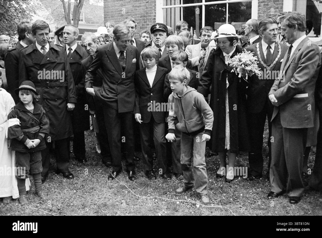 Queen's Day Veere Queen's Day Veere, Whizgle News, Dutch Desk, Paesi Bassi, 1950 - 2000 il 30-04-1981. Questi argomenti vengono visualizzati nell'immagine. In un ambiente all'aperto pieno di un mix di verde e architettura sobria, una folla di persone si riunisce, mostrando un arazzo di emozioni e interazioni. La scena presenta un gruppo eterogeneo, principalmente adulti in abbigliamento formale, tra cui abiti scuri completati da legami. Un ragazzo si distingue leggermente dal gruppo, vestito con un maglione casual di colore chiaro, la sua espressione porta un senso di urgenza o di angoscia, contrastando nettamente con gli adulti composti Foto Stock