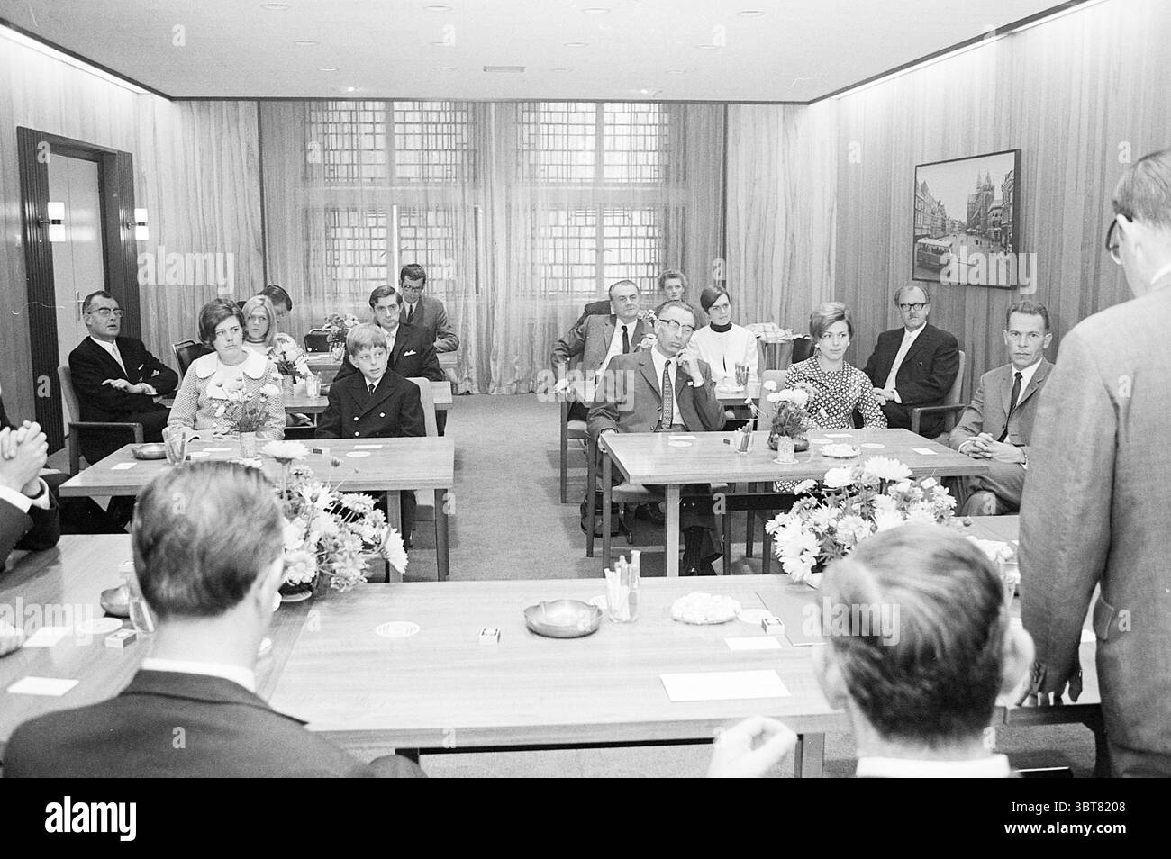 Reception Mr. De Ronde Noord Zuid Hollandse Vervoersmaatschappij N.V. NZ, Whizgle News, Dutch Desk, Paesi Bassi, 1950 - 2000 il 06-09-1969. Questi sono gli elementi dell'immagine. La scena cattura un luogo di ritrovo formale in una sala conferenze spaziosa e ben illuminata. Le pareti sono adornate con caldi pannelli di legno, che donano un'aria di raffinatezza all'ambiente. La luce naturale filtra attraverso le grandi finestre adornate da vetri decorativi, creando un'atmosfera morbida che esalta l'atmosfera generale della sala. Seduti intorno a diversi tavoli rettangolari vi sono numerosi partecipanti, uomini e donne, Foto Stock