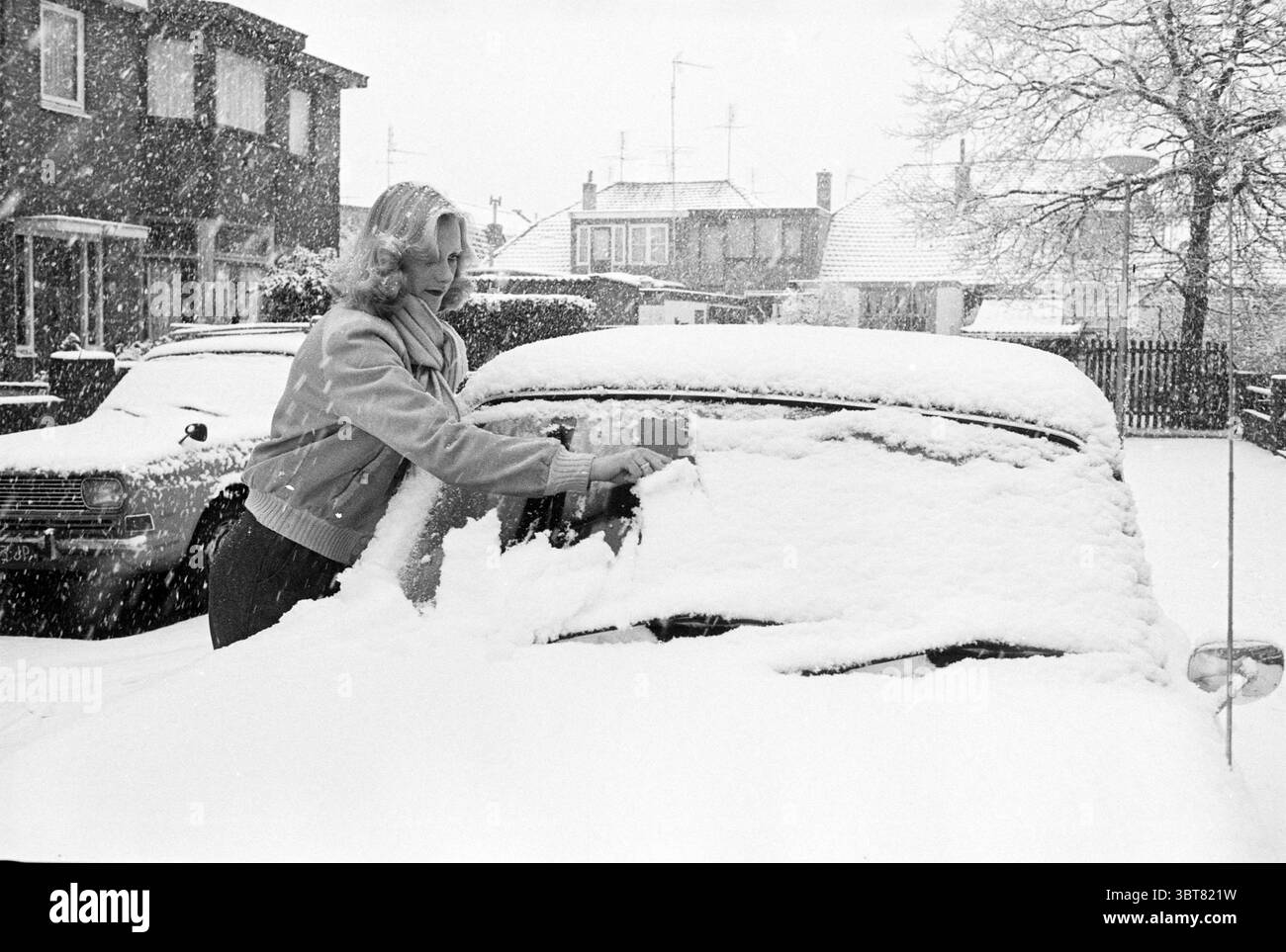 Sneeuwkiek Haarlem, Gd. Old Canal Snow Haarlem Gedempte Oude Gracht Paesi Bassi, Whizgle News, Dutch Desk, Paesi Bassi, 1950 - 2000 il 10-04-1978. Questi sono gli argomenti nell'immagine. In una scena invernale, una donna è impegnata nel compito di eliminare la neve da un'auto parcheggiata in una zona residenziale. Si trova accanto al veicolo, utilizzando un raschietto per rimuovere la neve spessa e polverosa che si accumula sul parabrezza. I suoi capelli sono modellati con onde morbide e indossa un cappotto accogliente e di colore chiaro che contrasta con il bianco intenso della neve. I dintorni sono dominati da una forte nevicata che crea un Foto Stock