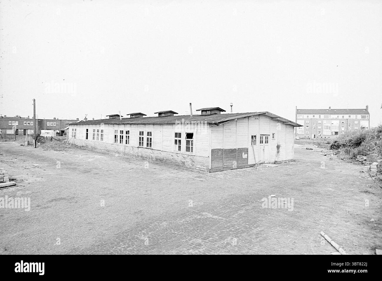 Est. Rotogravure Sleep-in Zandvoort Exterior, Whizgle News, Dutch Desk, Paesi Bassi, 1950 - 2000 il 19-05-1972. L'immagine include questi argomenti. La scena raffigura un ambiente desolato e scarsamente popolato. Al centro della composizione c'è una struttura in legno resistente agli agenti atmosferici, allungata e bassa al suolo, caratterizzata da una serie di finestre lungo la sua facciata, alcune delle quali appaiono incrinate o rotte. L'edificio ha un esterno macchiato, suggerendo anni di esposizione agli elementi. Intorno alla struttura è presente un terreno irregolare e ruvido, costituito principalmente da ciottoli, che aggiungono texture alla scena. La zona Foto Stock