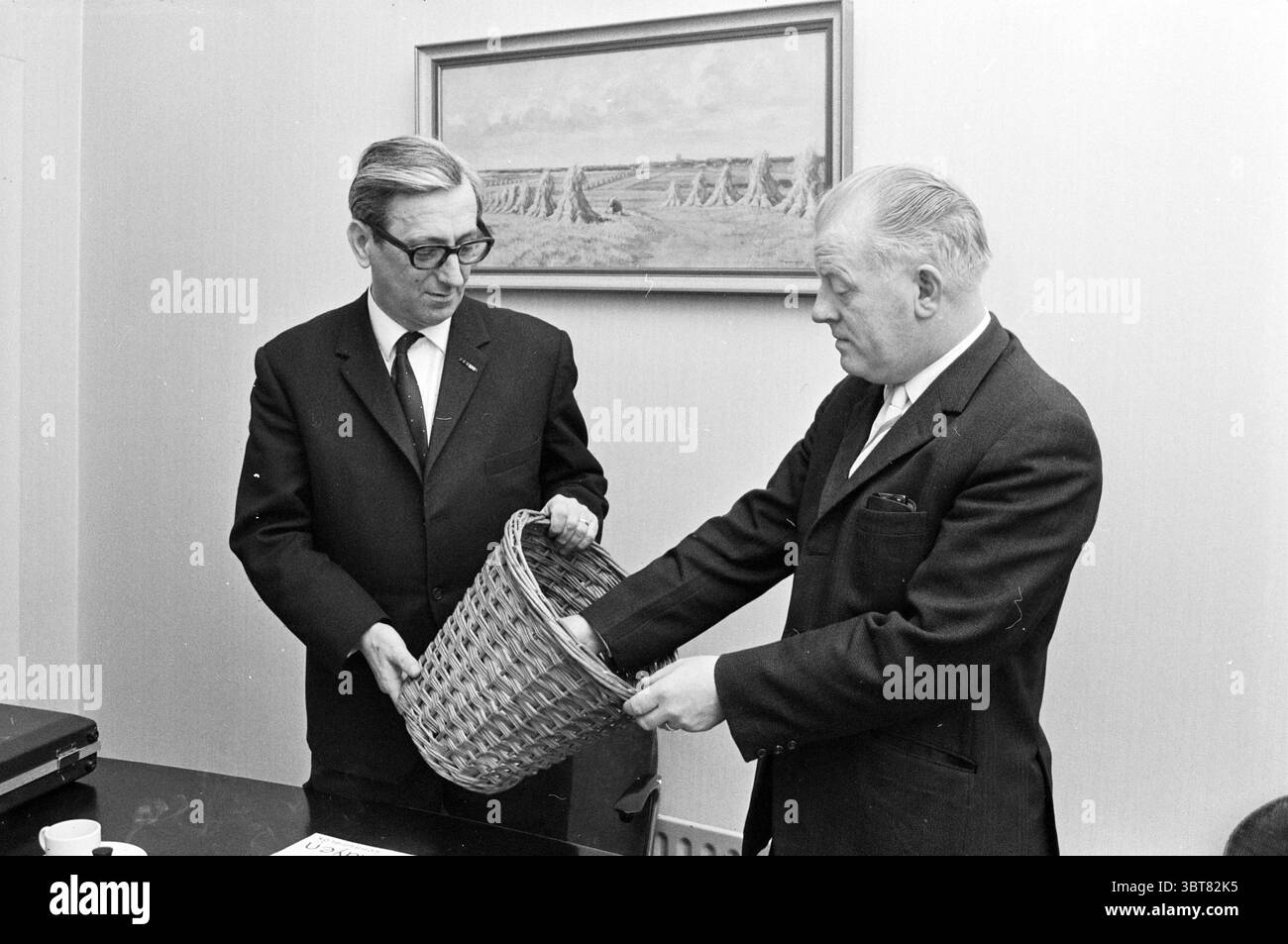 Draw AVG Lottery, Whizgle News, Dutch Desk, Paesi Bassi, 1950 - 2000 il 04-02-1970. Questi sono gli argomenti nell'immagine. In uno spazio interno dai toni chiari, due uomini sono impegnati in una conversazione. Un uomo, con occhiali e tuta scura, si erge a sinistra, reggendo un cesto in tessuto. La sua espressione sembra attenta mentre guarda verso l'altro uomo. Il secondo uomo, anch'esso vestito con un abito scuro ma senza occhiali, si erge a destra e sta entrando nel cesto, apparentemente esaminando il suo contenuto. Lo sfondo presenta un dipinto incorniciato che raffigura una serie di paesaggi stilizzati, principalmente in Foto Stock