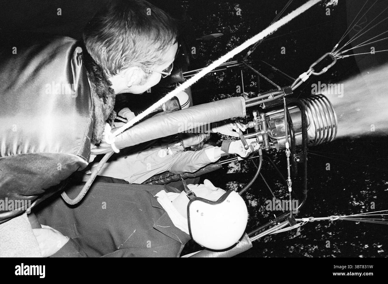 Sindaco Van Stam in mongolfiera Mayors Hoofddorp Municipal Council, Whizgle News, Dutch Desk, Paesi Bassi, 1950 - 2000 il 04-10-1980. Questi argomenti vengono visualizzati nell'immagine. In una scena dinamica, due persone sono impegnate nel funzionamento di una mongolfiera. Una persona si inclina in avanti, incentrata su un intricato sistema bruciatore, che emette un forte flusso di fiamma. I componenti metallici risplendono sotto il bagliore del fuoco, contrastando nettamente con l'ambiente circostante più scuro. L'altro individuo, seduto nel cesto, sembra calmo ma attento, indossando un casco di sicurezza come precauzione. La o Foto Stock