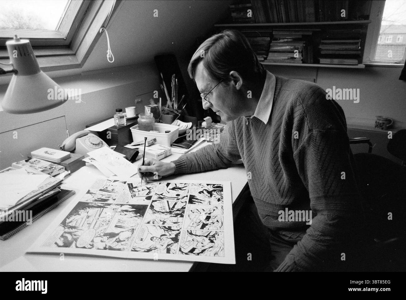 Fumettista Bert Bus, Whizgle News, Dutch Desk, Paesi Bassi, 1950 - 2000 il 19-11-1990. Questi argomenti vengono visualizzati nell'immagine. In un'accogliente area di lavoro con soffitti inclinati, un uomo è seduto a una scrivania, concentrandosi sull'attività che gli precede. Ha capelli corti e marrone chiaro e indossa occhiali, completati da un morbido maglione in maglia dalle tonalità tenui. La sua espressione trasmette la concentrazione mentre si muove leggermente in avanti, immergendo una penna nell'inchiostro e facendo dei tratti attenti su un grande foglio di carta sparso davanti a lui. La scrivania è piena di oggetti d'arte: Vasi pieni di pennelli, tavolozza, e. Foto Stock