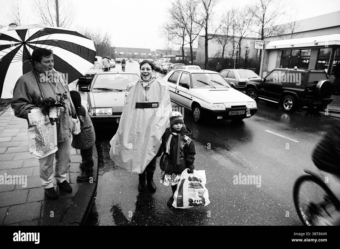 Padre del traffico, signora The Hope, Whizgle News, Dutch Desk, Paesi Bassi, 1950 - 2000 il 21-12-1995. L'immagine mostra questi argomenti. In una vivace scena urbana, gli individui si accoccolano sotto vari ombrelli in mezzo a una pioggia costante. Un adulto, con in mano un ombrello oversize a righe con audaci segmenti in bianco e nero, si trova vicino a un bambino piccolo che stringe una borsa, forse piena di dolci. L'adulto è vestito con un abbigliamento pratico e impermeabile, caratterizzato da toni tenui che contrastano delicatamente con l'abbigliamento vivace del bambino. Il bambino, sorridente, è indossato con un impermeabile luminoso che spicca di nuovo Foto Stock