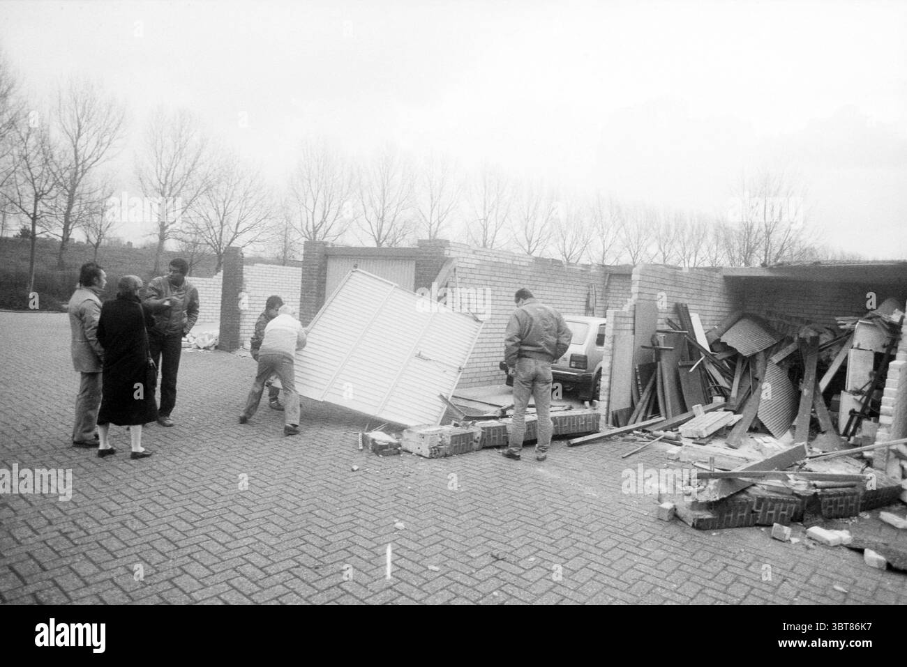 Storm Damage Haarlemmermeer various SEE folder Storm and Storm Damage, Whizgle News, Dutch Desk, Paesi Bassi, 1950 - 2000 on 26-01-1990. Questi sono gli argomenti nell'immagine. In un ambiente urbano cupo, un gruppo di cinque individui si riunisce intorno a mucchi di detriti, suggerendo una scena di recente demolizione o pulizia. Il paesaggio è caratterizzato da una serie di materiali irregolari impilati, tra cui tavole di legno e grandi lastre di materiale da costruzione, tutti sparpagliati a caso. Il terreno è pavimentato con mattoni grigi, che aggiungono un look strutturato ma non raffinato all'ambiente. La gente mi ha fidanzato Foto Stock