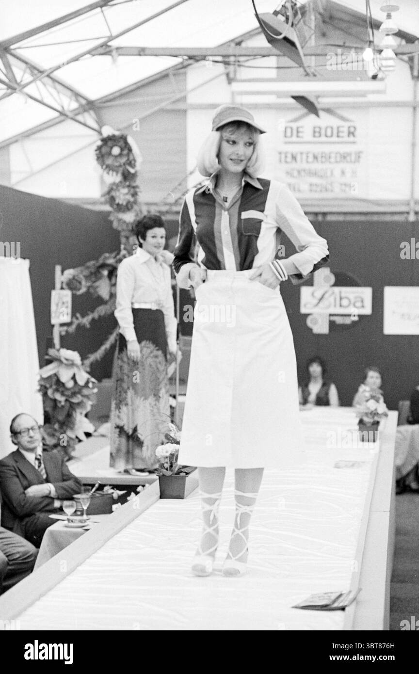 Sfilata di moda Liba Spring Fair, Whizgle News, Dutch Desk, Paesi Bassi, 1950 - 2000 il 10-03-1977. L'immagine mostra questi argomenti. La scena cattura una vivace sfilata di moda ambientata in una spaziosa arena al coperto, caratterizzata da una struttura simile a una tenda con un soffitto alto. Sulla passerella, un modello si muove con sicurezza, mostrando un outfit esclusivo che unisce elementi retrò e moderni. Indossa un top luminoso color block con strisce rosse, blu e bianche, abbinato a una gonna bianca a vita alta che scorre elegantemente intorno alle sue gambe. Il suo look è completato da un cappellino da baseball abbinato, Foto Stock