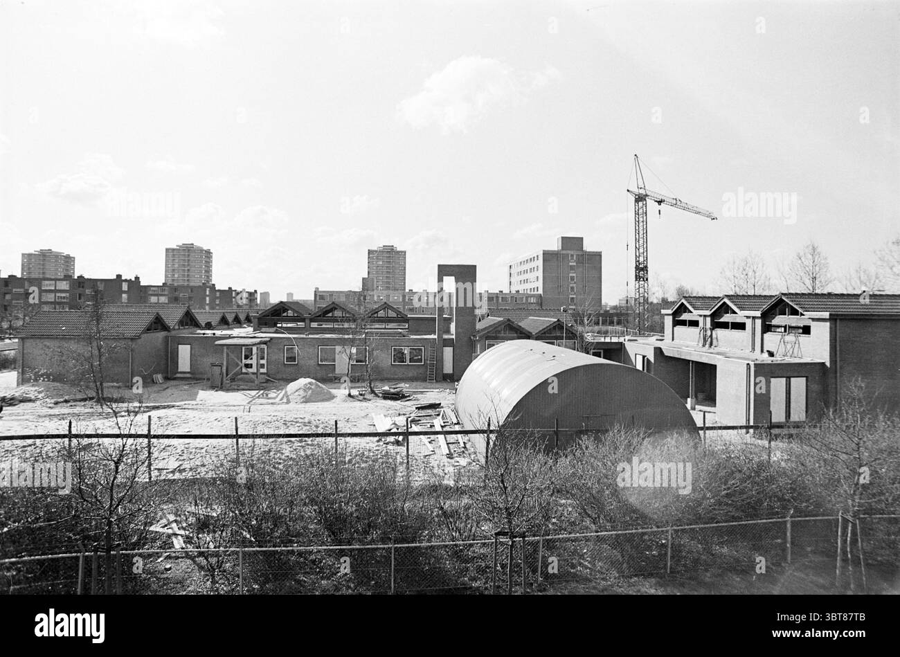 New van der Jagt School Jan Gijzenkade Haarlem + Old van der Jagt School on the Pr. Beatrixdreef in Parkwijk Schools Haarlem Prinses Beatrixdreef Paesi Bassi, Whizgle News, Dutch Desk, Paesi Bassi, 1950 - 2000 on 17-03-1975. Questi argomenti sono mostrati nell'immagine. La scena presenta un cantiere che mostra in modo prominente diverse strutture parzialmente costruite. Gli edifici hanno un mix di tetti piatti e leggermente inclinati, con numerose aperture per finestre che indicano le installazioni future. In primo piano, pile di materiali da costruzione (ghiaia, sabbia e utensili sparsi) suggeriscono un wor in corso Foto Stock