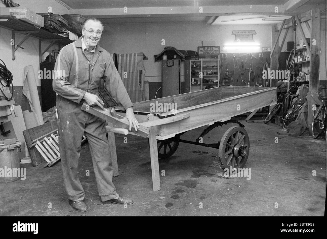 Restauro del vecchio carretto a mano di Lord Pel Restoration, Whizgle News, Dutch Desk, Paesi Bassi, 1950 - 2000 il 26-01-1988. Questi argomenti sono mostrati nell'immagine. In un'officina piena di attrezzi e materiali, un anziano si trova con orgoglio accanto a un carrello in legno parzialmente costruito. È vestito con un lavoro oscuro, che mostra un sorriso caratteristico che suggerisce un senso di realizzazione e nostalgia. Il carro, ancora nella sua forma grezza, ha una struttura di base in legno, che mette in risalto l'artigianalità della sua creazione. Intorno a lui, lo spazio è ingombrato da vari oggetti - scaffali lin Foto Stock