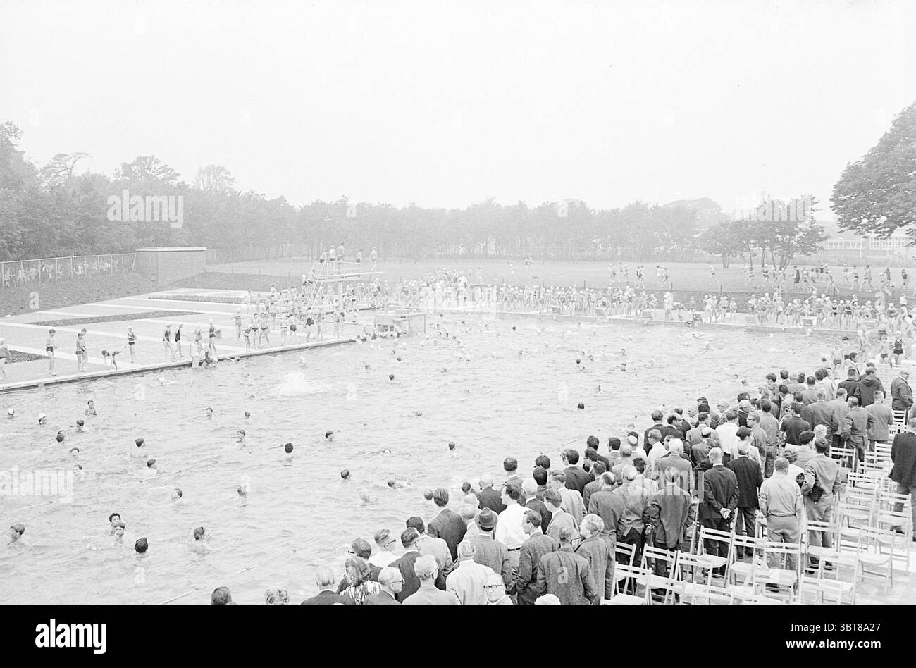 Apertura piscina Herenduinweg apertura aperture messo in uso IJmuiden Heerenduinweg Paesi Bassi, Whizgle News, Dutch Desk, Paesi Bassi, 1950 - 2000 il 17-06-1966. Questi argomenti sono mostrati nell'immagine. La scena si snoda in una vivace piscina all'aperto, piena di energia. La composizione cattura un grande raduno di persone, posizionato su entrambi i lati della piscina. Da un lato, una disposizione a più livelli di sedie in legno accoglie gli spettatori, che sembrano impegnati e animati, alcuni chiacchierano o puntano verso l'acqua. Nella piscina, una varietà di nuotatori portano in acqua, creando strappi Foto Stock