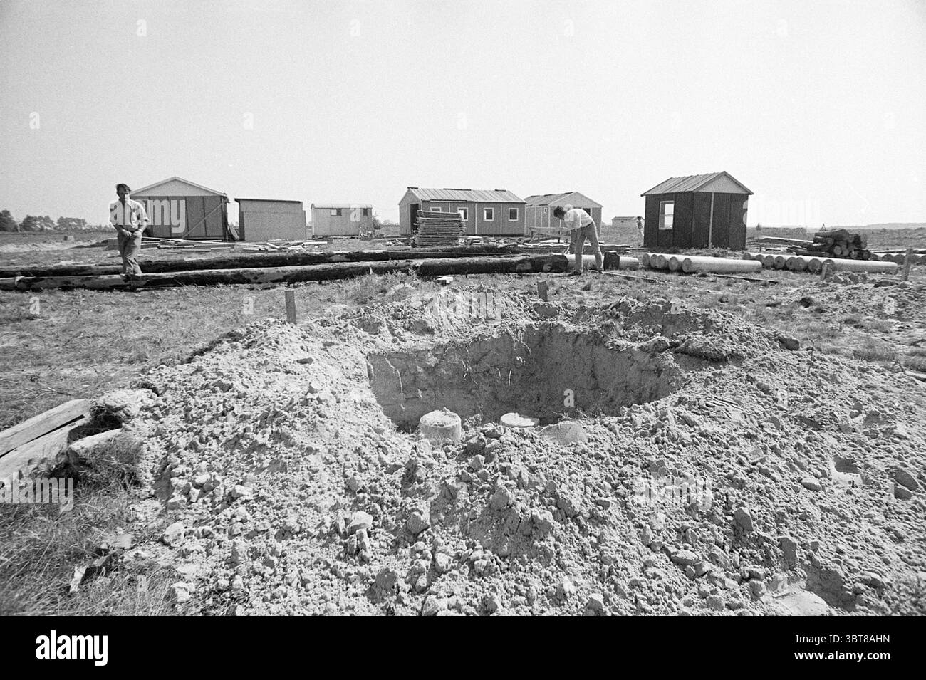 Preparativi per i lavori di costruzione, Whizgle News, Dutch Desk, Paesi Bassi, 1950 - 2000 il 28-08-1972. Questi argomenti sono mostrati nell'immagine. La scena cattura un cantiere rurale sotto un cielo chiaro e luminoso. In primo piano, una grande fossa circolare domina la composizione, la sua terra sabbiosa accumulata intorno ai bordi, suggerendo recenti scavi. Piccole pietre e detriti sono sparse vicino alla fossa, sottolineando i lavori in corso. Due figure sono visibili a sinistra e a destra, entrambe vestite con abiti da lavoro tipici del lavoro manuale, tra cui camicie e pantaloni robusti. Sembrano concentrati Foto Stock