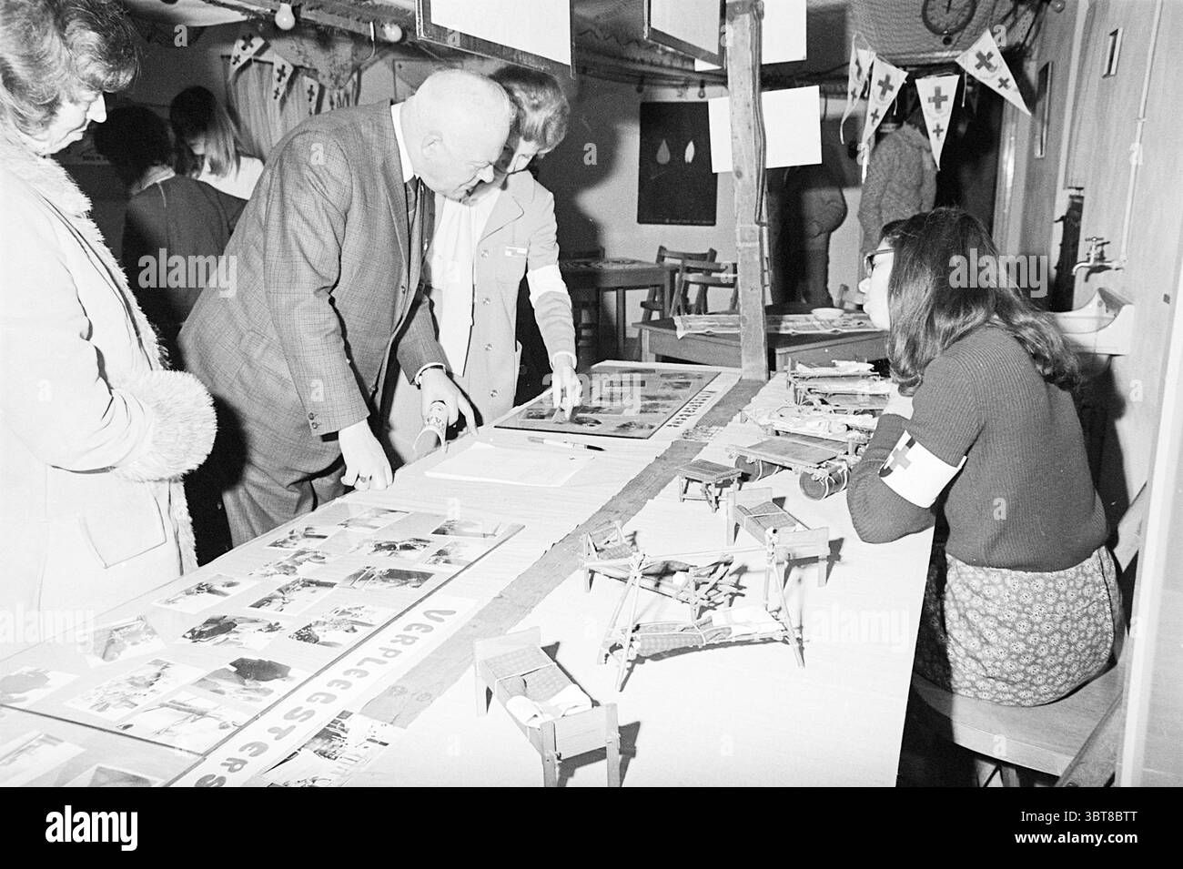 Croce Rossa Giovanile Velsen Croce Rossa Croce Rossa ospedale, Whizgle News, Dutch Desk, Paesi Bassi, 1950 - 2000 il 09-01-1971. Questi sono gli elementi dell'immagine. In un vivace ambiente interno, un lungo tavolo si estende in primo piano, coperto da una serie di fotografie e piccoli modelli. La scena è vivace, piena di persone impegnate in conversazione ed esame. Ci sono quattro individui in primo piano. A sinistra, una donna che indossa un cappotto invernale sta in piedi, osservando attentamente mentre si piega leggermente in avanti. Accanto a lei, un uomo illustre con un abito oscuro e gesti di cravatta animati, Foto Stock