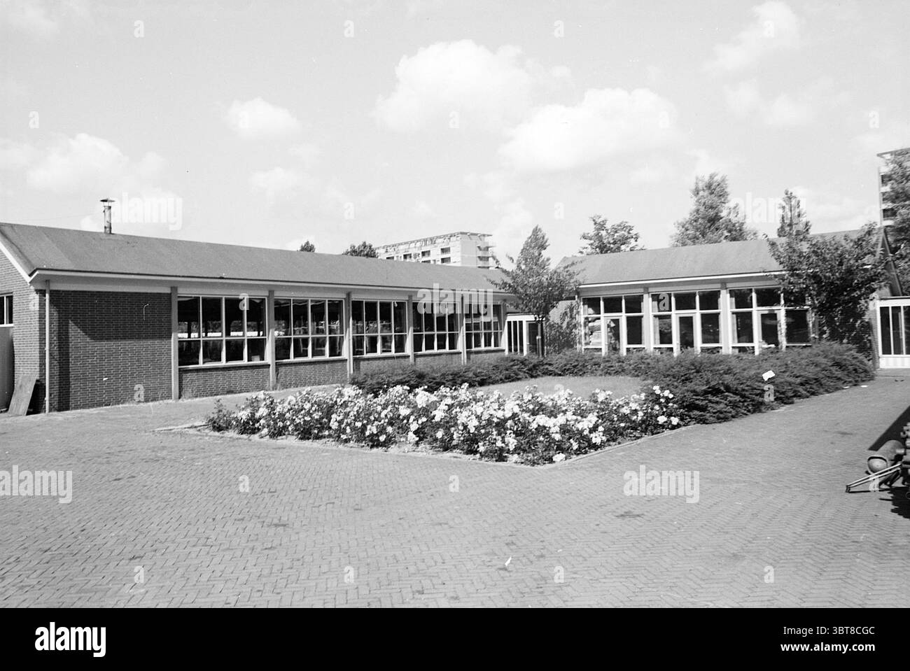 Edificio scolastico., Whizgle News, Dutch Desk, Paesi Bassi, 1950 - 2000. L'immagine mostra questi argomenti. La scena raffigura una spaziosa area esterna caratterizzata da distinte strutture architettoniche. Due edifici adiacenti con tetti piani sono disegnati in modo semplice, i loro esterni in mattoni principalmente in toni tenui. Le grandi finestre dominano le facciate, permettendo alla luce naturale di inondare gli interni e dando un senso di apertura. Di fronte agli edifici, un giardino ben curato esplode con una varietà di piante fiorite, formando un vibrante contrasto con il verde circostante. Il flowe Foto Stock