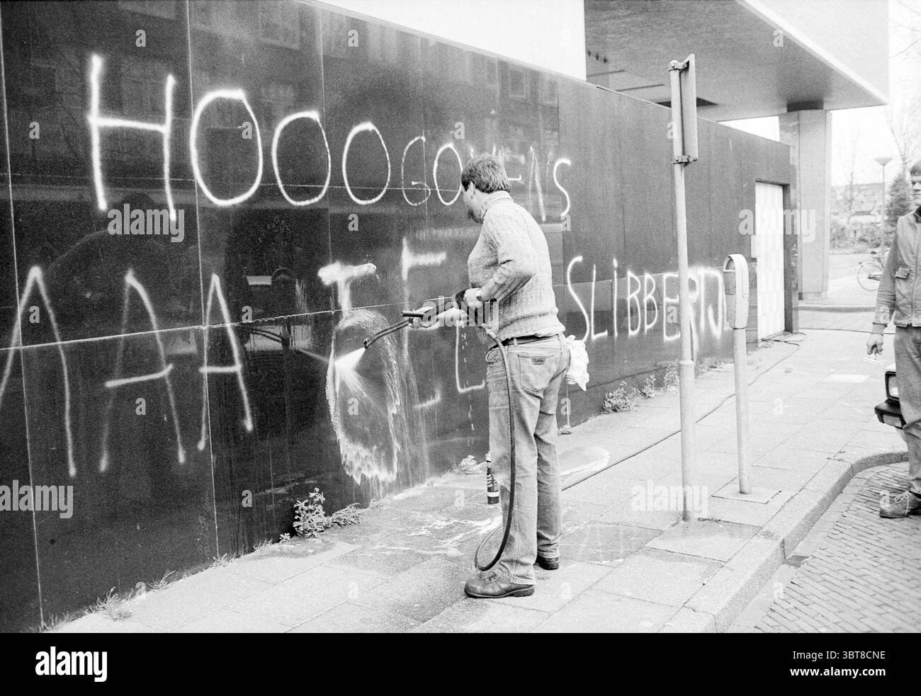 Il municipio di IJmuiden ha sfrontato i testi dei piani costieri IJmuiden Nederland, Whizgle News, Dutch Desk, Paesi Bassi, 1950 - 2000 il 19-04-1989. Questi sono gli elementi dell'immagine. La scena cattura un angolo di strada in città dove una persona è impegnata a pulire un grande muro scuro. La parete è parzialmente oscurata dai graffiti, con lettere audaci dipinte a spruzzo bianche che contrastano nettamente con la superficie nera profonda. La pulizia individuale è vestita con un abbigliamento casual, che consiste in una camicia di colore chiaro e jeans, e utilizza una lavatrice ad alta pressione, che invia flussi d'acqua sul Foto Stock