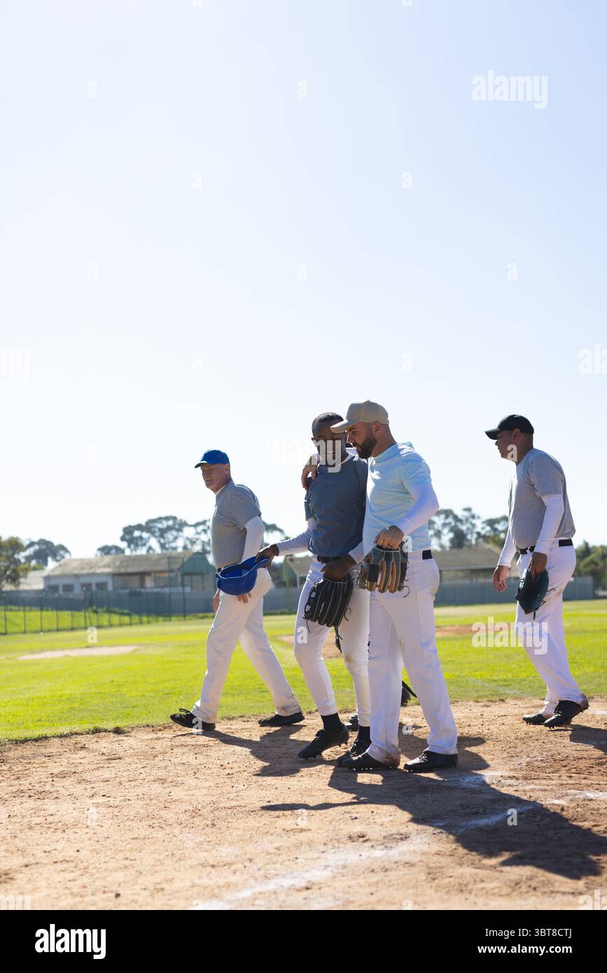 Diversi compagni di squadra maschili che camminano attraverso lo sporco del campo da baseball tenendo guanti e berretti Foto Stock