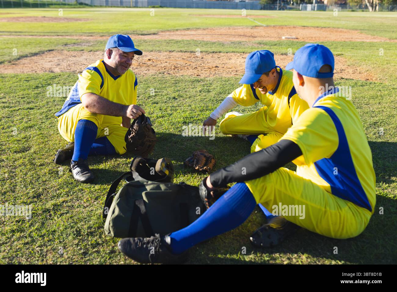 I compagni di squadra selezionano guanti e chiacchierano con maglie gialle blu sull'erba del campo da baseball Foto Stock