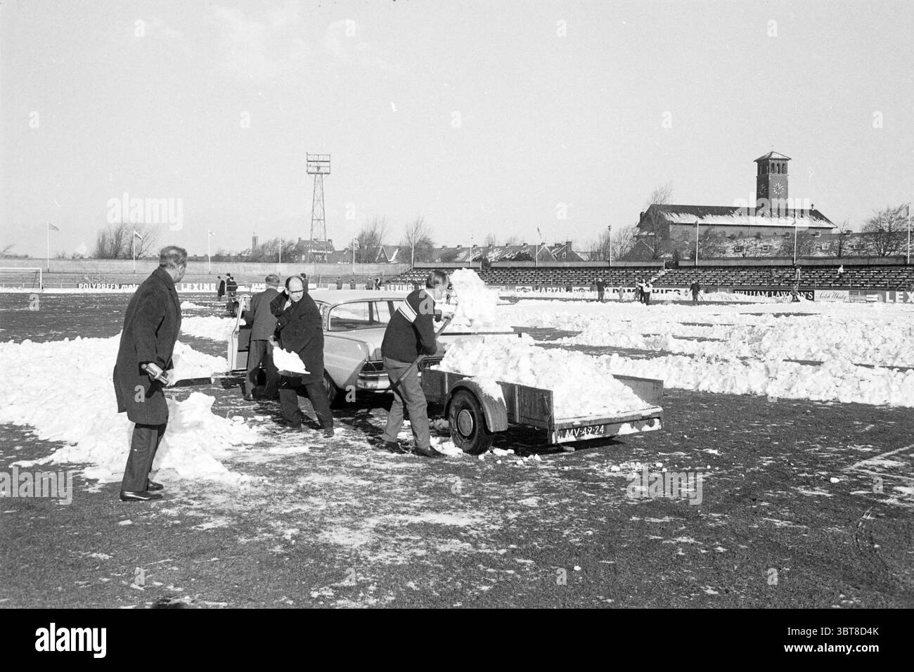 Disinnevamento in Haarlem Grounds Football Haarlem Work, Whizgle News, Dutch Desk, Paesi Bassi, 1950 - 2000 il 04-02-1968. Questi sono gli elementi dell'immagine. La scena cattura un paesaggio invernale, dominato da un grande campo aperto coperto da uno spesso strato di neve. Il cielo sopra è un grigio pallido e coperto, che suggerisce un'atmosfera fredda. In primo piano, diversi individui sono attivamente impegnati nella rimozione della neve, creando macchie chiare sul campo e i loro respiri visibili nell'aria ghiacciata. I lavoratori sono vestiti con cappotti e cappelli scuri, in contrasto con la neve bianca brillante. Qualche ar Foto Stock