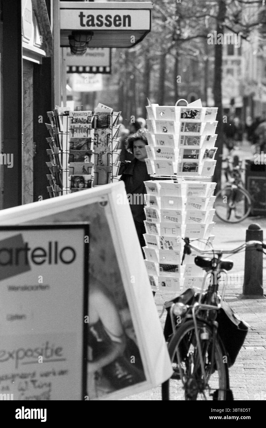 Scopri il materiale per vetrine in Street Haarlem Grote Houtstraat Paesi Bassi, Whizgle News, Dutch Desk, Paesi Bassi, 1950 - 2000 il 23-03-1993. Questi argomenti sono mostrati nell'immagine. La scena raffigura un vivace ambiente stradale, con particolare attenzione a un marciapiede affollato, fiancheggiato da vari oggetti e attività. In primo piano, gli scaffali espongono un assortimento di volantini e opuscoli, suggerendo la presenza di negozi o gallerie nelle vicinanze. La tavolozza dei colori è prevalentemente monocromatica e presenta varie sfumature di grigio, che trasmettono un'atmosfera senza tempo e un po' nostalgica. Più indietro, una persona sta in piedi sl Foto Stock
