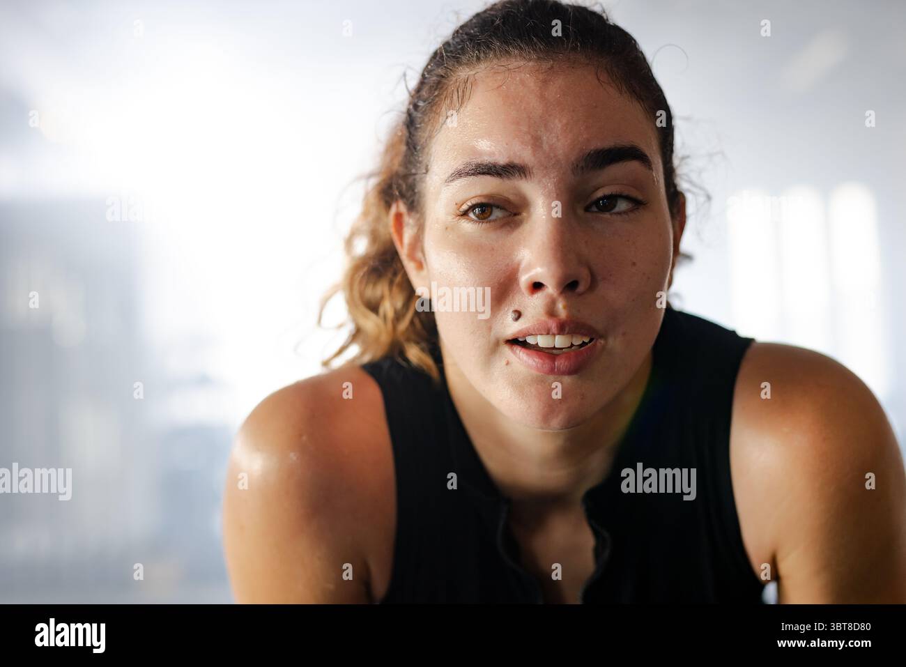 Atleta che si piega in avanti dopo l'allenamento tra le macchine da palestra sfocate e indossa canotta nera Foto Stock