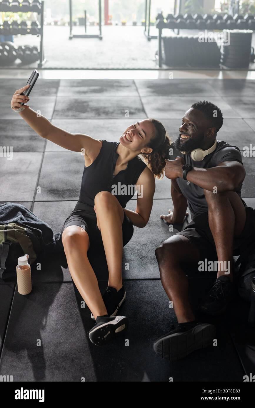 Diversi partner di allenamento seduti sul tappetino della palestra che scattano selfie con smartphone, cuffie e bottiglia d'acqua Foto Stock