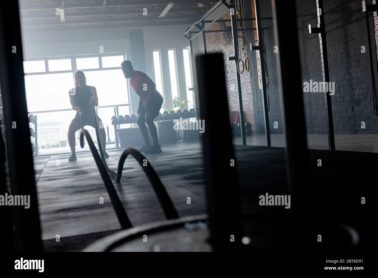 Atleta bianca che tira corde da combattimento mentre allenatrice maschile nera in palestra con piatti Foto Stock