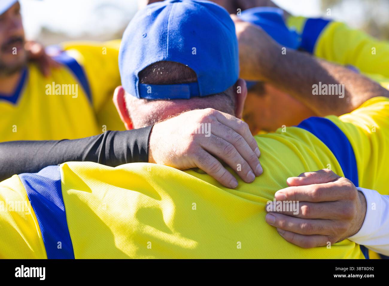 Compagni di squadra maschi senior che si accoccolano prima della partita sul campo da baseball indossando maglie gialle e cappellino blu Foto Stock