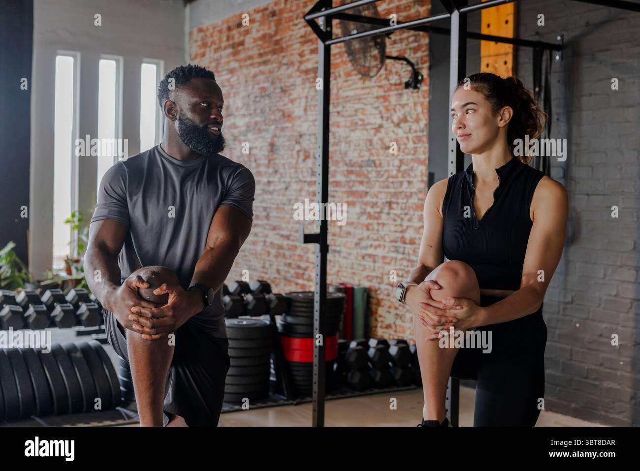 Diversi partner di allenamento che si allungano in palestra su tappetini in gomma con cremagliera per manubri e squat rack Foto Stock