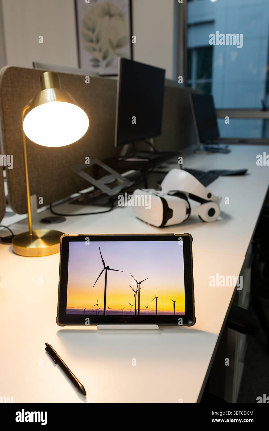 Tablet in custodia protettiva che mostra le turbine eoliche al tramonto sulla scrivania con lampada e visore VR Foto Stock