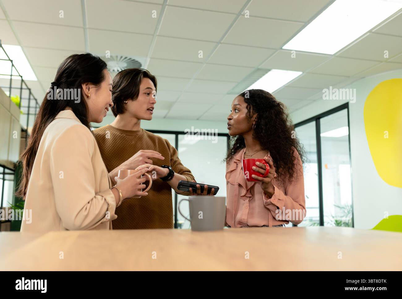 Diversi colleghi che chiacchierano al piano di lavoro in legno nell'angolo cottura dell'ufficio con tazze e smartphone Foto Stock