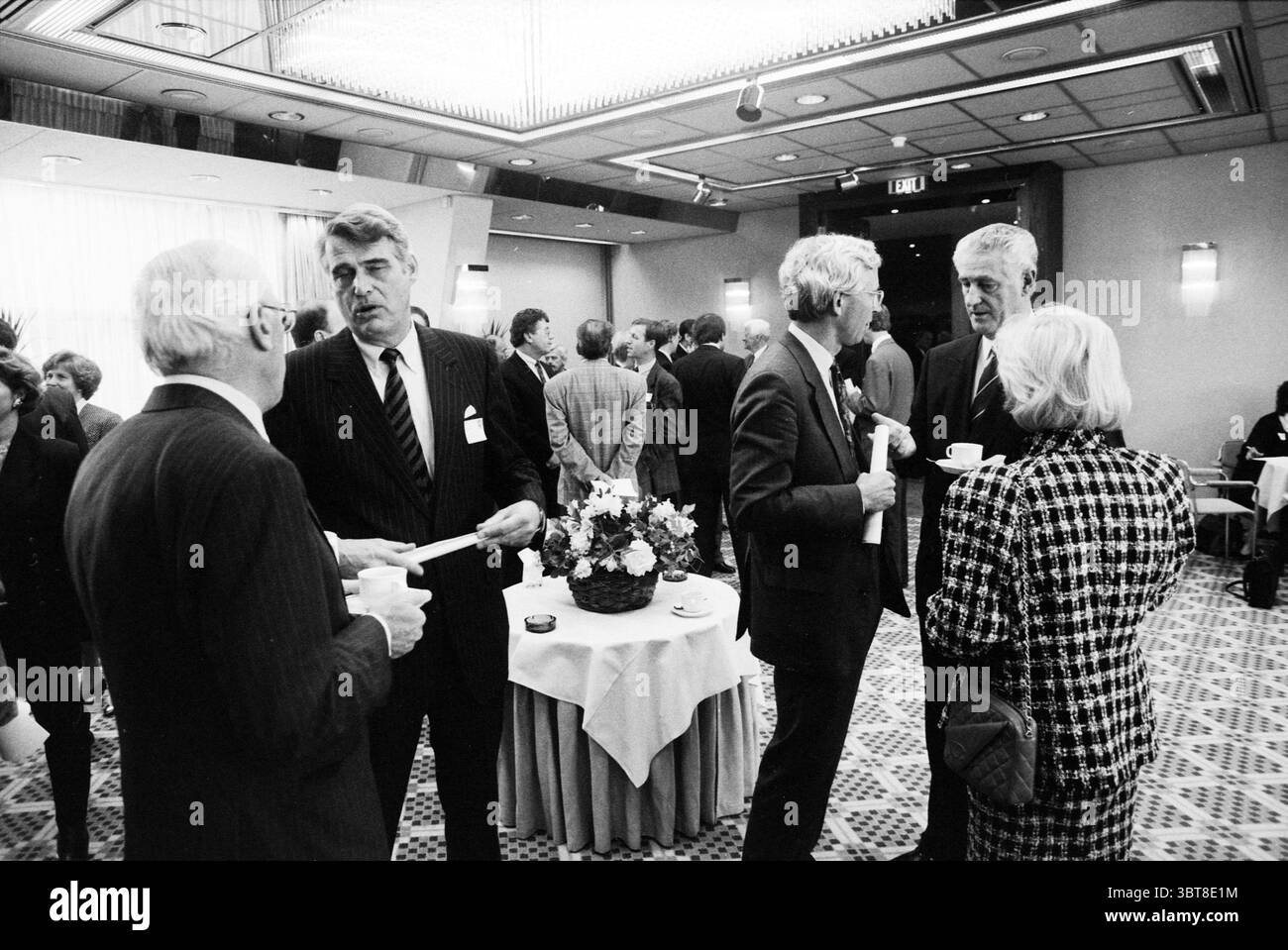 M. Schröder Schiphol Hilton Chamber of Commerce Schiphol, Whizgle News, Dutch Desk, Paesi Bassi, 1950 - 2000 il 22-04-1991. Questi sono gli argomenti nell'immagine. La scena si sviluppa in un ambiente interno sofisticato, probabilmente una sala per conferenze o riunioni. L'atmosfera e' animata da un gruppo di persone ben vestite impegnate in conversazione. Gli uomini sono prevalentemente vestiti con abiti scuri, con alcune variazioni nei colori cravatta, mentre le donne mostrano un mix di stili, tra cui motivi e texture classici. Una grande disposizione floreale occupa il centro di una tavola rotonda, disegnando attentio Foto Stock