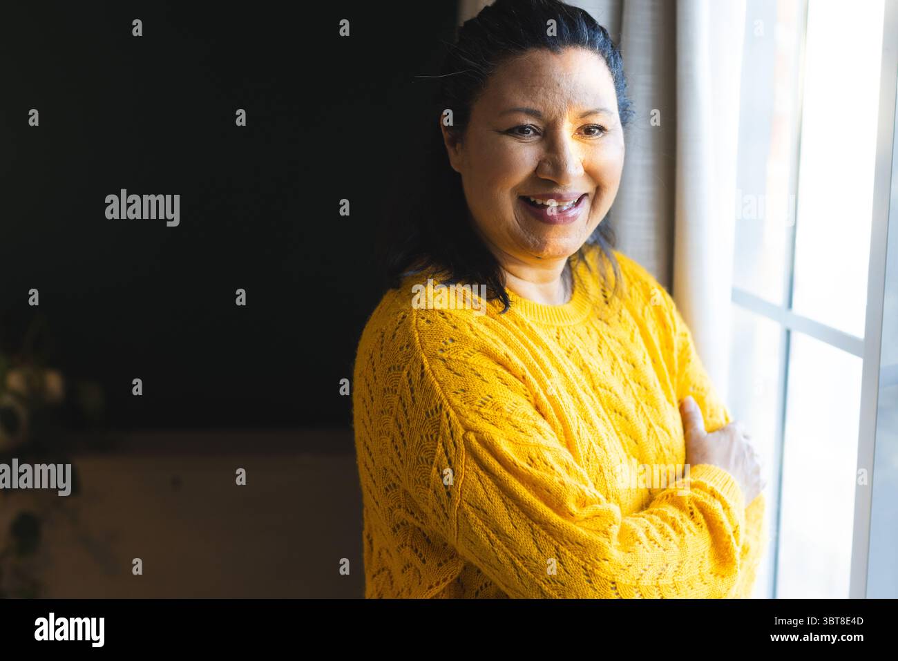 Donna in piedi a casa accanto alla cornice della finestra in maglione giallo sorridente e guardando all'esterno, copia spazio Foto Stock