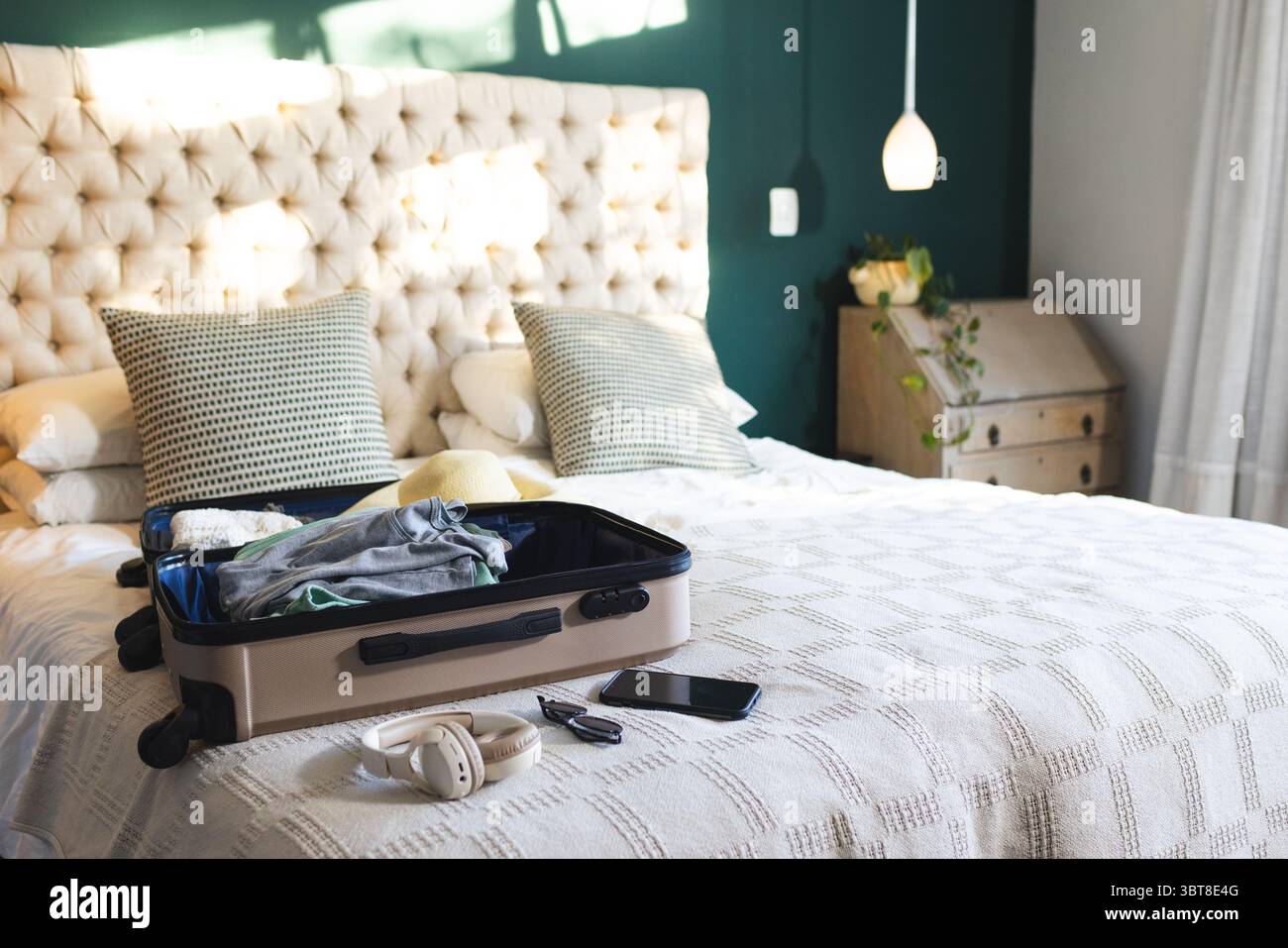 Valigia aperta beige appoggiata su un letto fatto con cuscini a motivi geometrici e cuffie contro il muro di tè Foto Stock