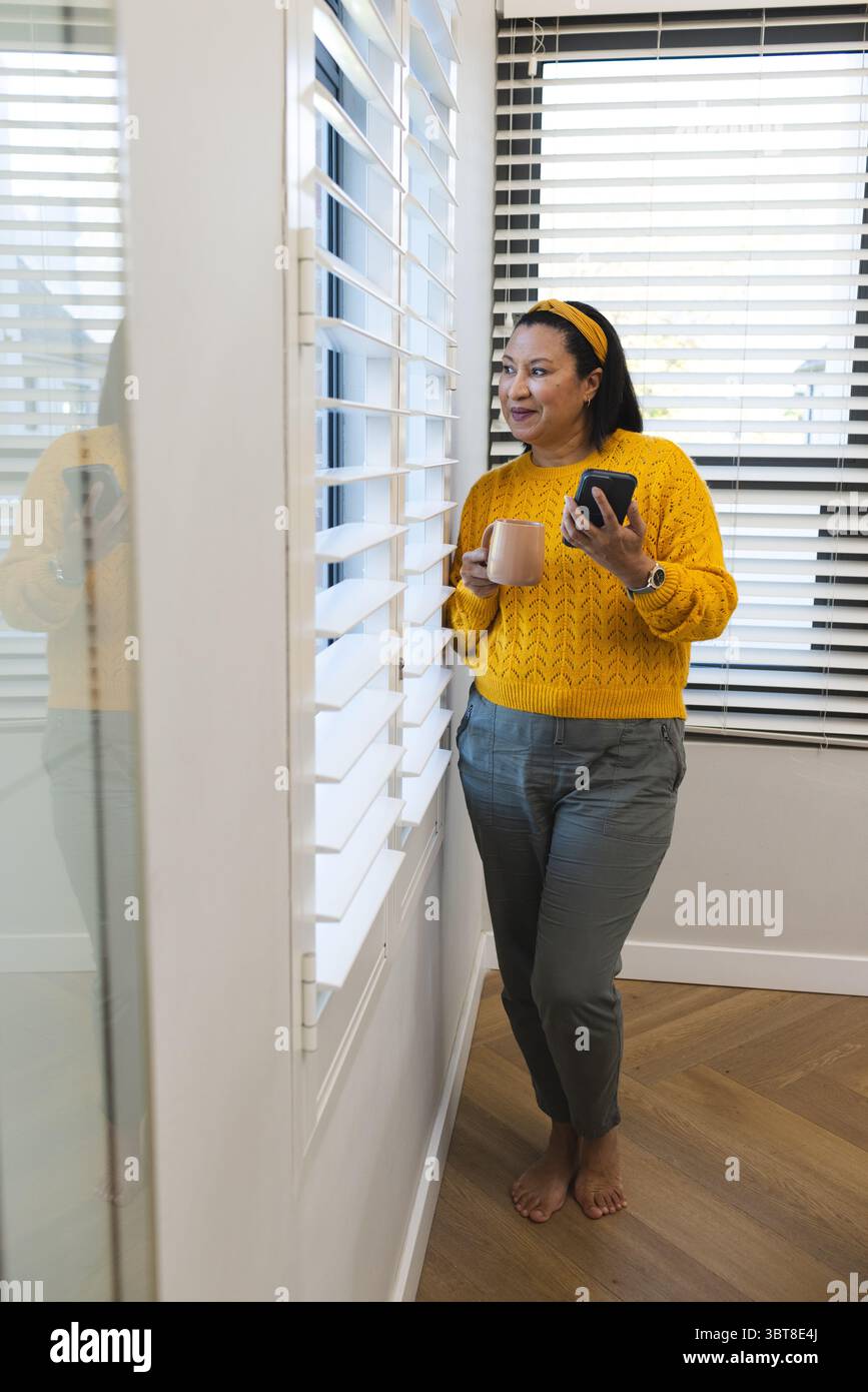 Donna a piedi nudi su pavimenti in legno vicino a una finestra con persiane, che tiene in mano tazze e smartphone, spazio per fotocopie Foto Stock