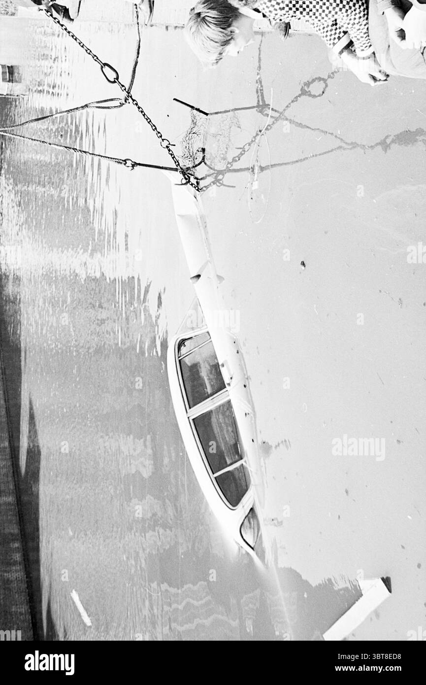 Un'auto caduta in acqua nel Burgwal viene sollevata Haarlem nei Paesi Bassi ., Whizgle News, Dutch Desk, Paesi Bassi, 1950 - 2000. Questi sono gli elementi dell'immagine. La scena presenta un ambiente oscuro e monocromatico dominato da varie sfumature di grigio. In prima linea, un'auto sommersa è parzialmente visibile, il suo profilo emerge appena sopra la superficie dell'acqua. I finestrini del veicolo sono offuscati, a indicare le profondità torbide sottostanti. Nelle vicinanze, un giovane individuo, vestito con un motivo a scacchi, si appoggia sul bordo dell'acqua, apparentemente affascinato dalla vista. L'angolo della loro B. Foto Stock