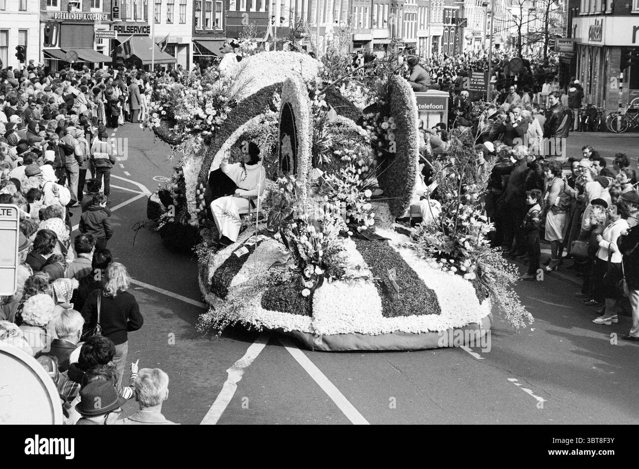 Sfilata di fiori sfilata di fiori mostra di fiori, Whizgle News, Dutch Desk, Paesi Bassi, 1950 - 2000 il 24-04-1982. L'immagine contiene questi argomenti. La scena è vivace e vivace, piena di una grande folla che si riunisce da vicino lungo i lati di una strada. Le persone sono vestite in vari stili, riflettendo un mix di abbigliamento casual e festivo. Alcuni sorridono, catturano l'emozione dell'evento, mentre altri sono impegnati in conversazioni animate o puntano verso la sfilata. Al centro dell'attenzione c'è un grande galleggiante adornato da un'elaborata esposizione di fiori e fogliame. Il fl Foto Stock