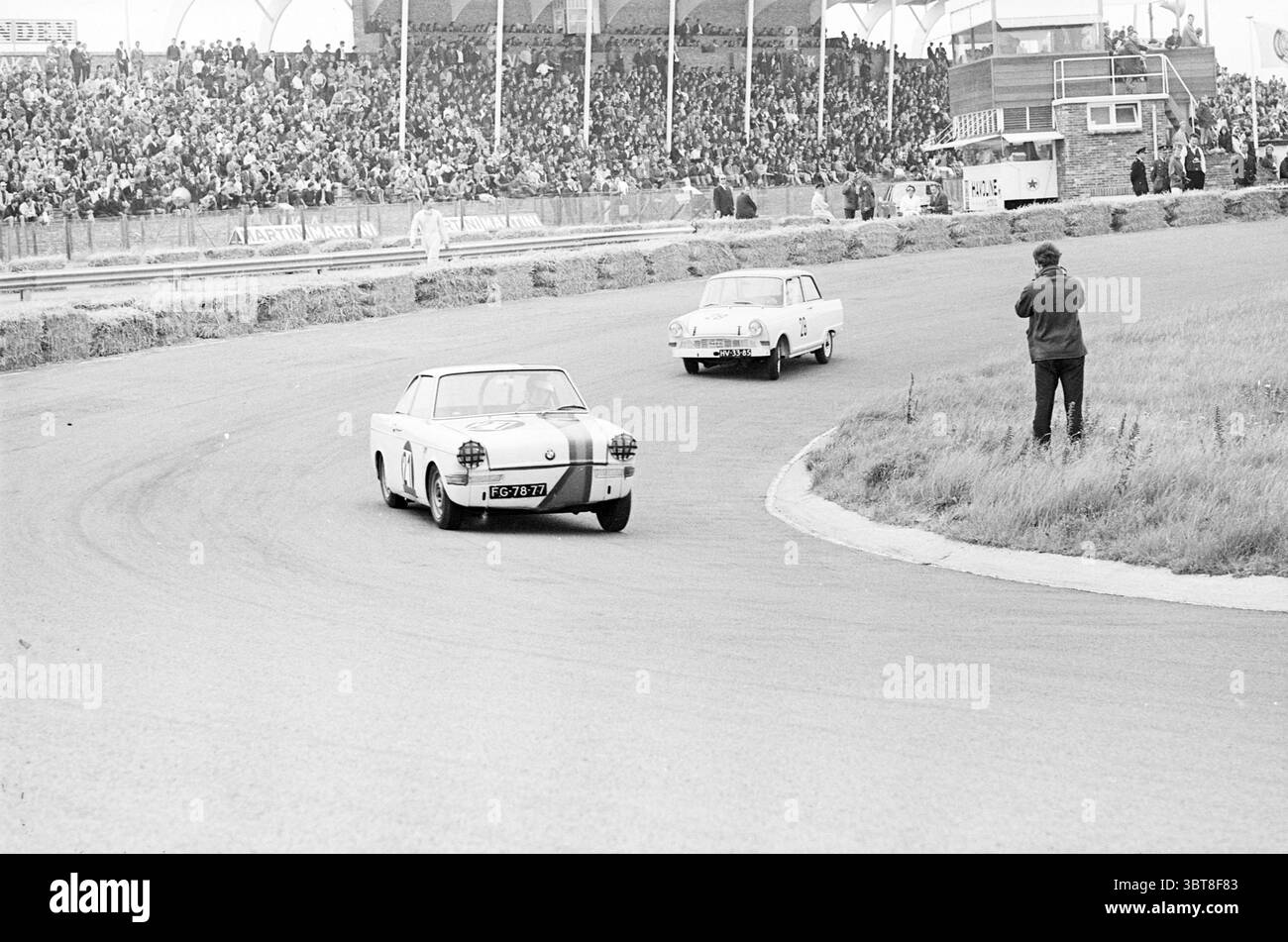 Gare automobilistiche sul circuito di Zandvoort Zandvoort, Whizgle News, Dutch Desk, Paesi Bassi, 1950 - 2000 il 18-07-1964. Questi sono gli argomenti nell'immagine. La scena raffigura un evento sportivo dinamico su un circuito, pieno di emozioni e anticipazioni. In primo piano, due auto d'epoca percorrono la curva del tracciato. Un'auto, dal design elegante in bianco, sembra essere all'avanguardia, mentre la seconda, leggermente dietro, è un veicolo più arrotondato, che mostra un'era diversa del design automobilistico. Entrambe le auto hanno un fascino nostalgico, che incarna l'estetica classica delle corse. Intorno alla pista, un'enthusia Foto Stock