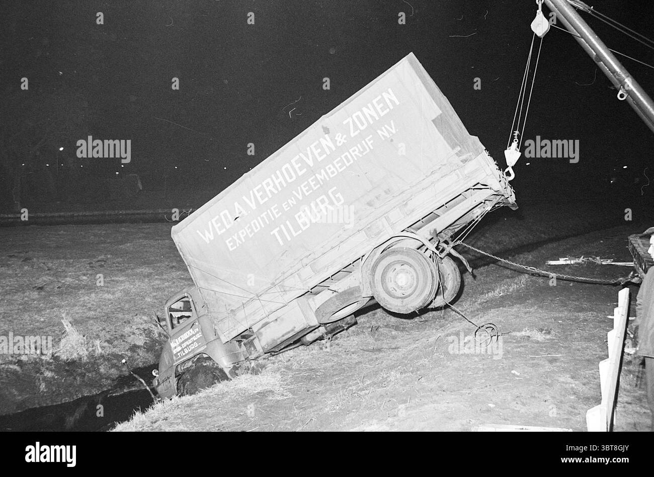 Un camion caduto in acqua viene tirato fuori dal fosso, Whizgle News, Dutch Desk, Paesi Bassi, 1950 - 2000 il 22-03-1965. Questi sono gli argomenti nell'immagine. La scena raffigura un'ambientazione notturna con un camion rovesciato sul bordo di un fosso. Il veicolo, che ha visto giorni migliori, è posizionato ad angolo, con le ruote posteriori sollevate e la parte anteriore parzialmente sommersa nel fosso. La sua superficie è accentuata da un esterno intemprato, leggermente danneggiato, che suggerisce anni di utilizzo. Lo schema di colori include toni attenuati come il grigio opaco e sfumature, che si aggiungono al sommo generale Foto Stock