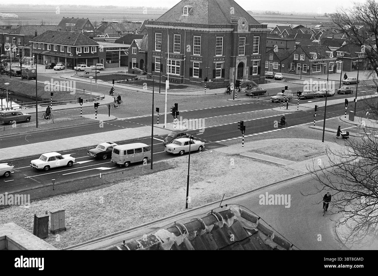 Incrocio Hoofdstraat in Hoofddorp Road Construction Road Planting Hoofddorp Hoofdstraat Paesi Bassi, Whizgle News, Dutch Desk, Paesi Bassi, 1950 - 2000 il 17-04-1964. Questi sono gli elementi dell'immagine. La scena cattura un vivace incrocio urbano con una miscela di stili architettonici. Centrale per la vista c'è un edificio in mattoni, probabilmente un'ex struttura municipale, caratterizzato da un tetto a picco e grandi finestre che suggeriscono il suo significato storico. Attorno ad esso si trovano edifici residenziali e commerciali, caratterizzati da caratteristici palazzi e altezze variabili, Foto Stock