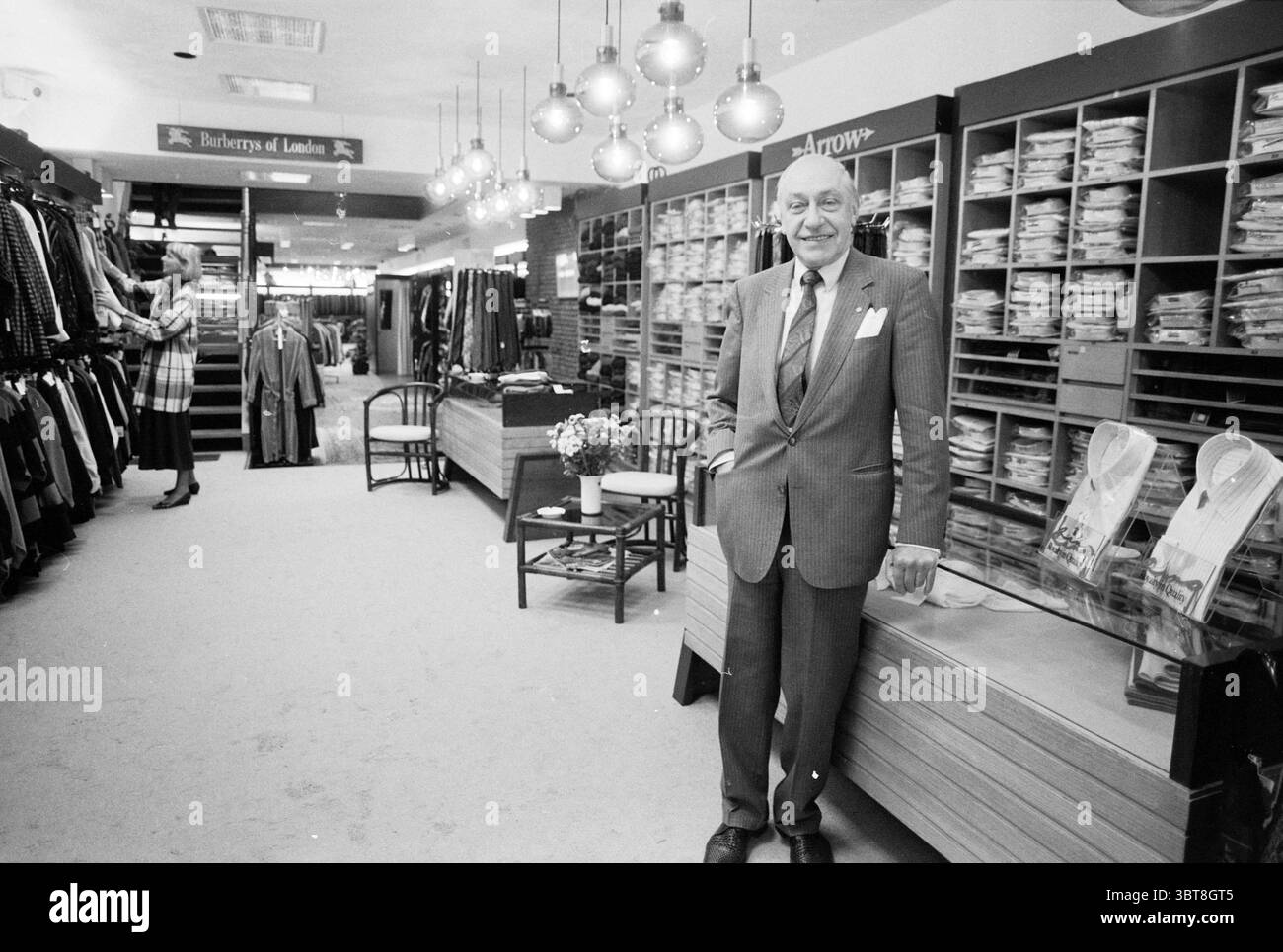 Mr. Da Kimade nei negozi e chioschi Haarlem Paesi Bassi, Whizgle News, Dutch Desk, Paesi Bassi, 1950 - 2000 il 12-09-1986. L'immagine include questi argomenti. La scena raffigura un ampio negozio di abbigliamento con un'atmosfera organizzata e invitante. A sinistra, una donna ben vestita sta sfogliando una cremagliera di vestiti, la sua figura parzialmente oscurata dalla mostra, suggerendo un senso di curiosità e interesse. In primo piano c'è un uomo di mezza età con una tuta, che trasuda fascino e fiducia. Ha un sorriso caldo e sembra accogliente, con le mani casualmente poste davanti Foto Stock