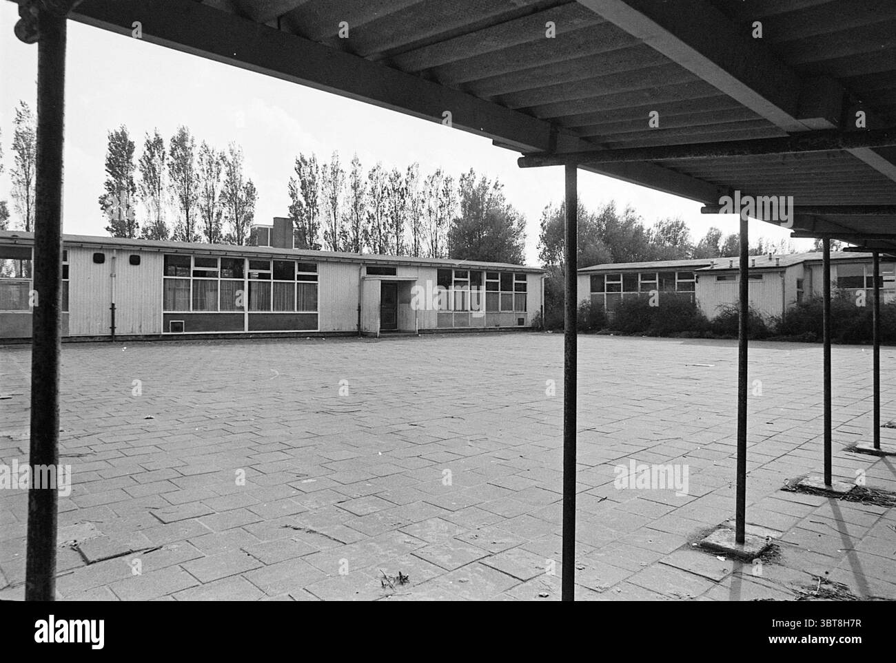 Panoramica Leao School Brink Beverwijk Schools, Whizgle News, Dutch Desk, Paesi Bassi, 1950 - 2000 il 17-10-1976. L'immagine contiene questi argomenti. La scena presenta un cortile desolato e desolato circondato da una serie di bassi edifici rettangolari. L'architettura appare utilitaria, con tetti piatti e grandi finestre, alcune delle quali sono parzialmente oscurate dalla mancanza di manutenzione. Le pareti degli edifici sono dipinte in toni tenui, somiglianti al bianco sbiadito o al grigio chiaro, che si fondono con l'atmosfera complessivamente cupa dell'ambiente. In primo piano, un baldacchino di travi scure si estende su t Foto Stock