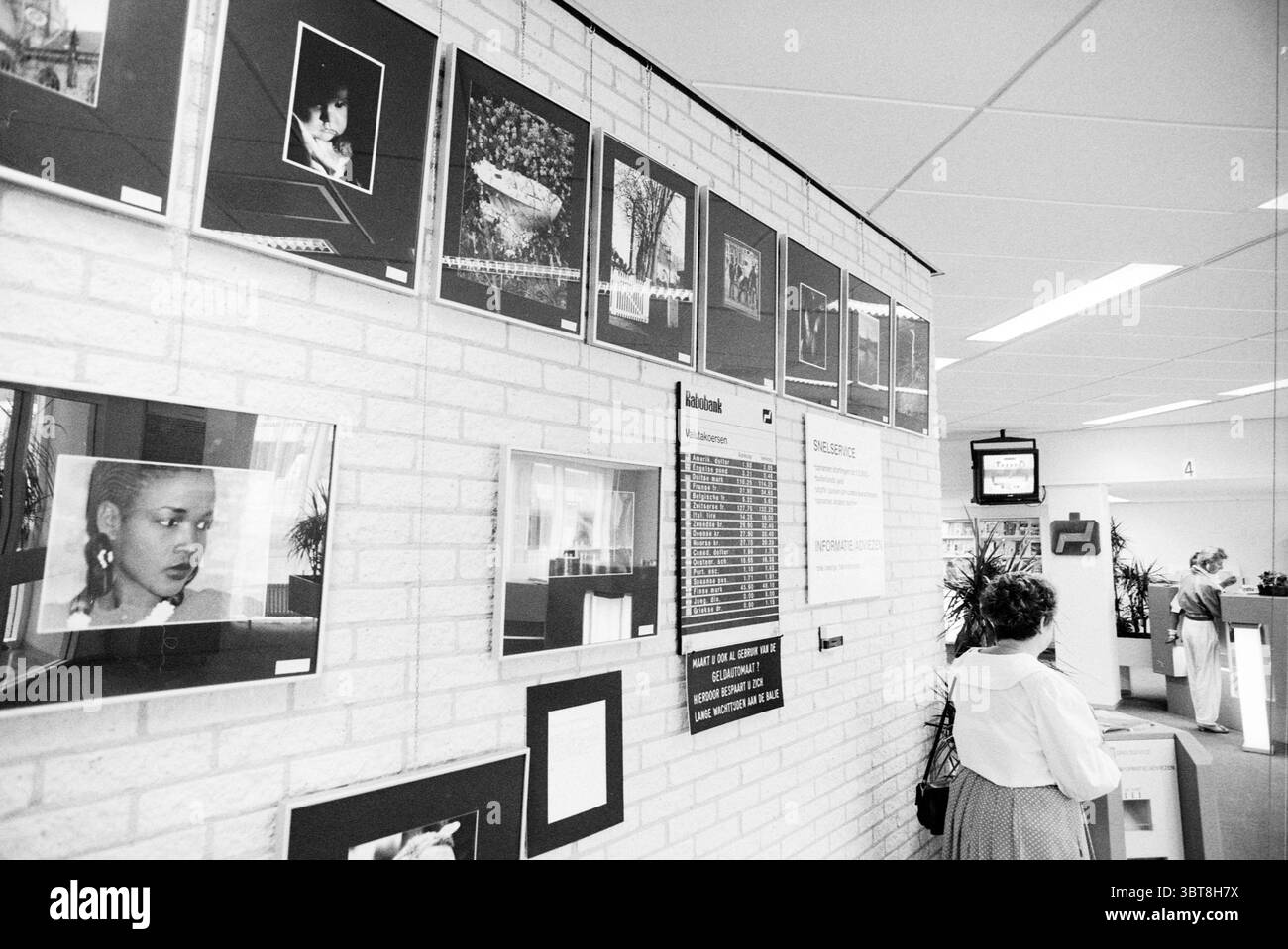 Mostra fotografica Rabobank IJmuiden IJmuiden Paesi Bassi, Whizgle News, Dutch Desk, Paesi Bassi, 1950 - 2000 il 24-07-1991. Questi argomenti vengono visualizzati nell'immagine. La scena è ambientata in un ambiente interno spazioso e ben illuminato, probabilmente in una galleria o in uno spazio pubblico. Una parete è dominata da una serie di fotografie incorniciate in bianco e nero disposte in modo ordinato in una griglia. Le immagini variano a seconda del soggetto, con ritratti, paesaggi e composizioni astratte, creando un arazzo di contrasti visivamente ricco. In primo piano, una persona sta con le spalle allo spettatore, di fronte al muro di Foto Stock
