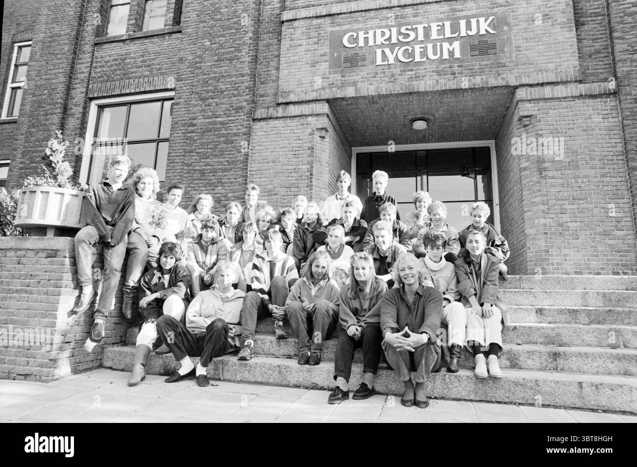 Prima Chr. Lyceum class Photo Schools, Whizgle News, Dutch Desk, Paesi Bassi, 1950 - 2000 on 29-10-1986. Questi sono gli argomenti nell'immagine. In un ambiente esterno ben strutturato, un grande gruppo di individui si riunisce sui gradini di un prominente edificio in mattoni. L'architettura presenta forme robuste e rettangolari, con alte finestre e una facciata simmetrica. L'entrata dell'edificio e' invitante, incorniciata da un'ampia scala, su cui gli individui sono seduti e in piedi. Le persone in questa scena mostrano un mix di espressioni e posture, suggerendo un mix di personalità. Bambini e teena Foto Stock