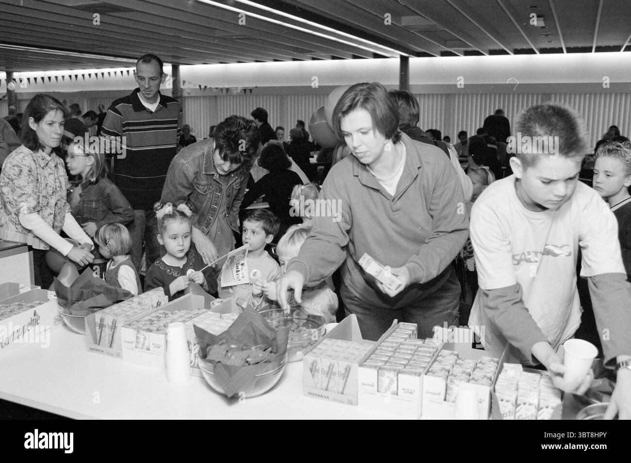 Saint Nicholas a Fluor Haarlem Paesi Bassi, Whizgle News, Dutch Desk, Paesi Bassi, 1950 - 2000 il 30-11-2001. Questi argomenti vengono visualizzati nell'immagine. In un vivace ambiente interno, numerose persone sono impegnate in varie attività. L'atmosfera e' vivace, piena di chiacchiere e il suono dei bambini che ridono. Lo spazio è pieno di lunghi tavoli carichi di oggetti colorati. Un tavolo prominente è coperto in contenitori ben disposti, probabilmente riempiti di spuntini o bevande. Nelle vicinanze, un gruppo di bambini, alcuni con capelli luminosi e abiti vivaci, sembrano impazienti ed eccitati, raggruppandosi Foto Stock