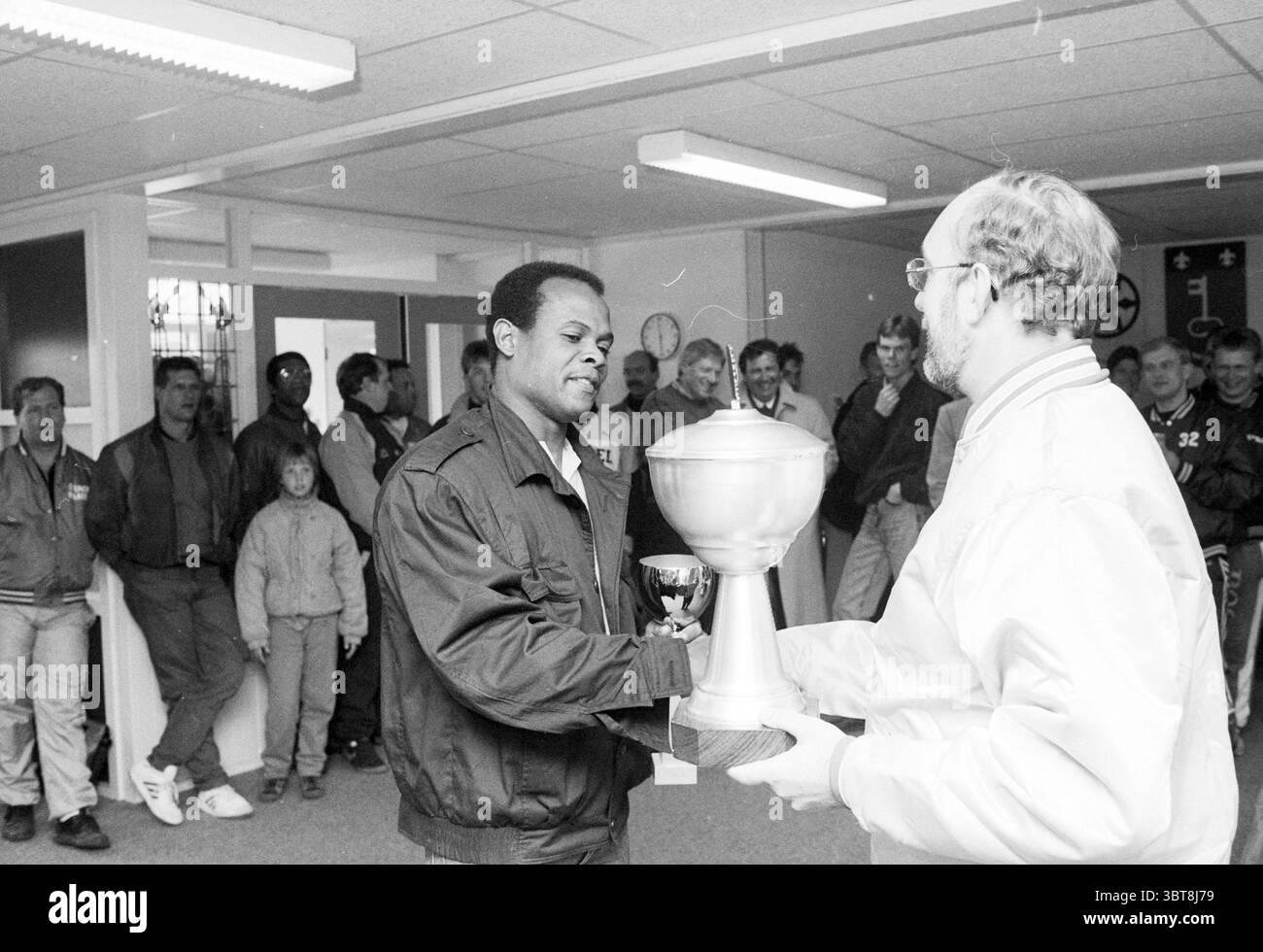 Cerimonia di premiazione Nicols Baseball Tournament Baseball Haarlem Nicols, Whizgle News, Dutch Desk, Paesi Bassi, 1950 - 2000 il 10-04-1988. L'immagine contiene questi argomenti. In un interno spazioso e ben illuminato, un gruppo di persone si riunisce, creando un senso di comunità e festa. Le pareti sono dipinte in un colore neutro, migliorando la luminosa illuminazione superiore. Al centro della scena, un uomo con pelle scura è impegnato in un momento di riconoscimento, accettando un grande trofeo da un altro individuo. Il trofeo ha una finitura metallica lucida e le sue dimensioni impressionanti suggeriscono un risultato significativo. TH Foto Stock