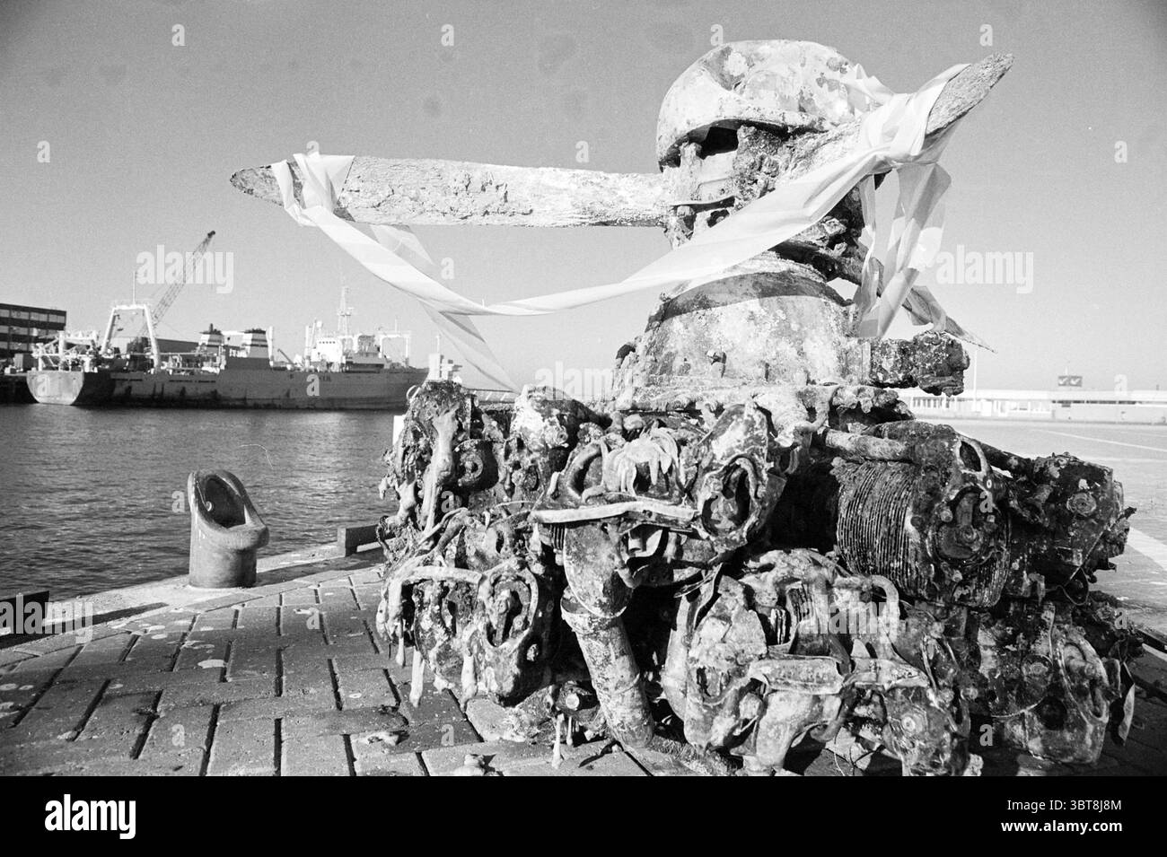 Motore aereo della seconda guerra mondiale, Whizgle News, Dutch Desk, Paesi Bassi, 1950 - 2000 il 13-10-1994. Questi sono gli argomenti nell'immagine. La scena presenta un grande oggetto intemprato che sembra essere l'ancora di una nave, fortemente corroso e ricoperto di ruggine. Questo tassello è situato su un molo, con una superficie in cemento testurizzato che mostra crepe e usura, suggerendo anni di esposizione a condizioni marine difficili. Intorno all'ancora, ci sono numerosi barnacli che si aggrappano alla sua superficie, evidenziando il suo tempo sommerso dall'acqua. Dietro l'ancora, il porto si estende, con diverse barche che saltano dolcemente Foto Stock