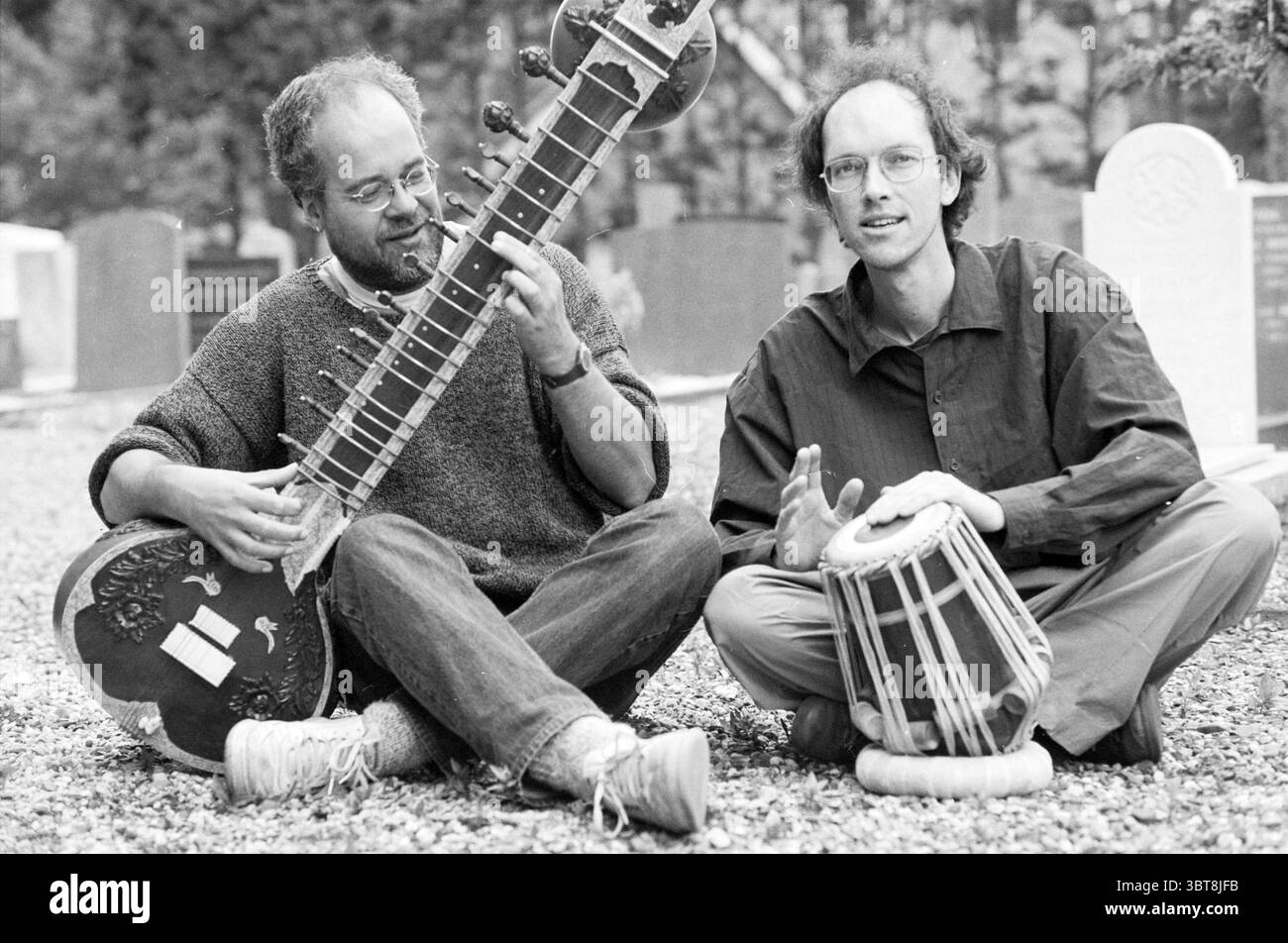 Proprietario Crazy Days, Whizgle News, Dutch Desk, Paesi Bassi, 1950 - 2000 il 14-07-1991. Questi sono gli argomenti nell'immagine. In un ambiente tranquillo all'aperto, due musicisti sono seduti vicino su un sentiero di ghiaia, circondati da un'atmosfera di tranquillità. Lo sfondo presenta file di lapidi intemperie, suggerendo che questo raduno si svolga in un cimitero, dando un'atmosfera contemplativa alla scena. Un musicista è impegnato con un grande sitar, uno strumento dal design complesso con un collo grazioso e ricurvo e un corpo risonante che riflette la maestria della musica tradizionale indiana. La loro f Foto Stock